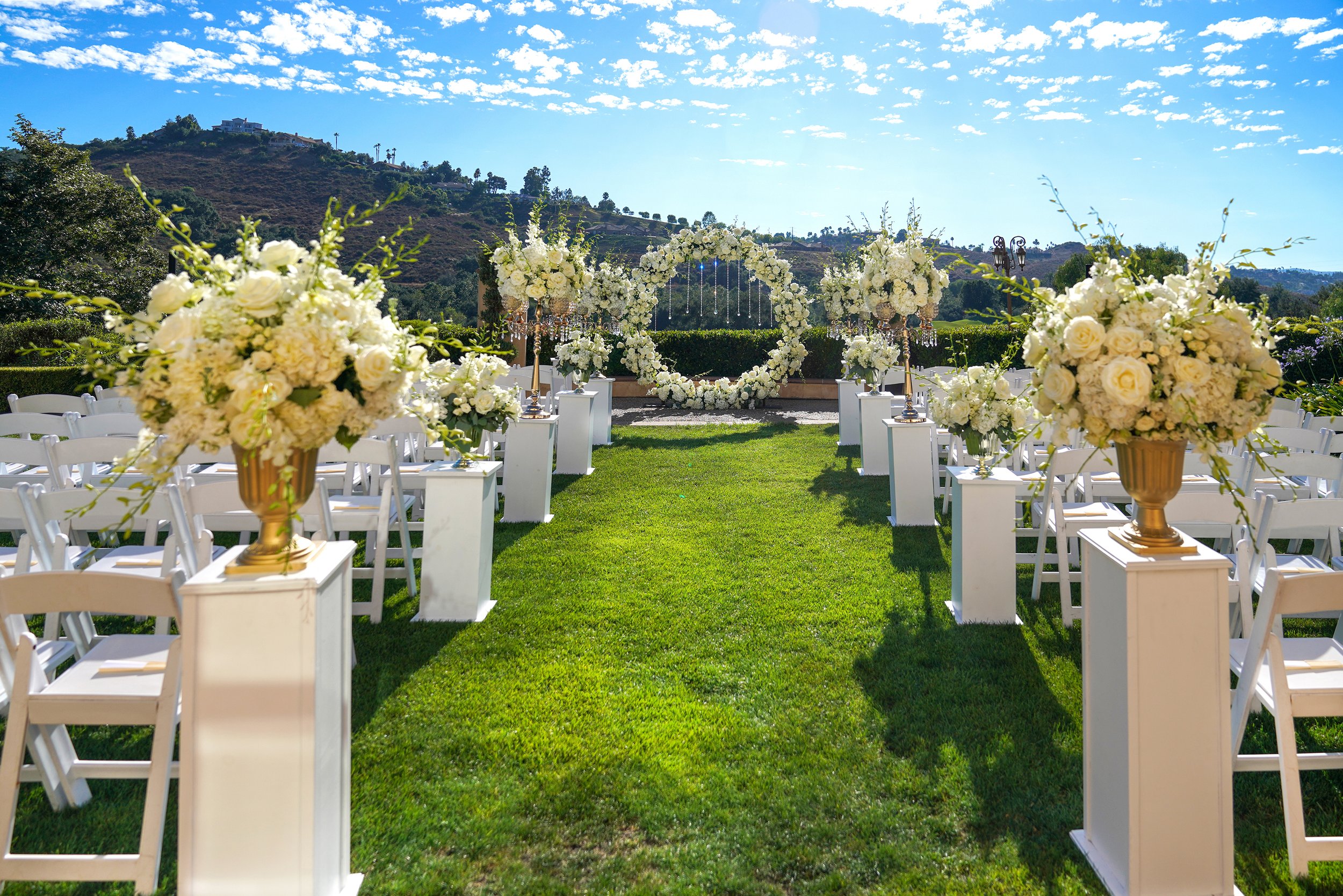 An outdoor wedding ceremony setup on green grass featuring white chairs lining an aisle toward a floral wedding circle or arch, with large white floral arrangements on gold urns, and a backdrop of hills under a bright blue sky.