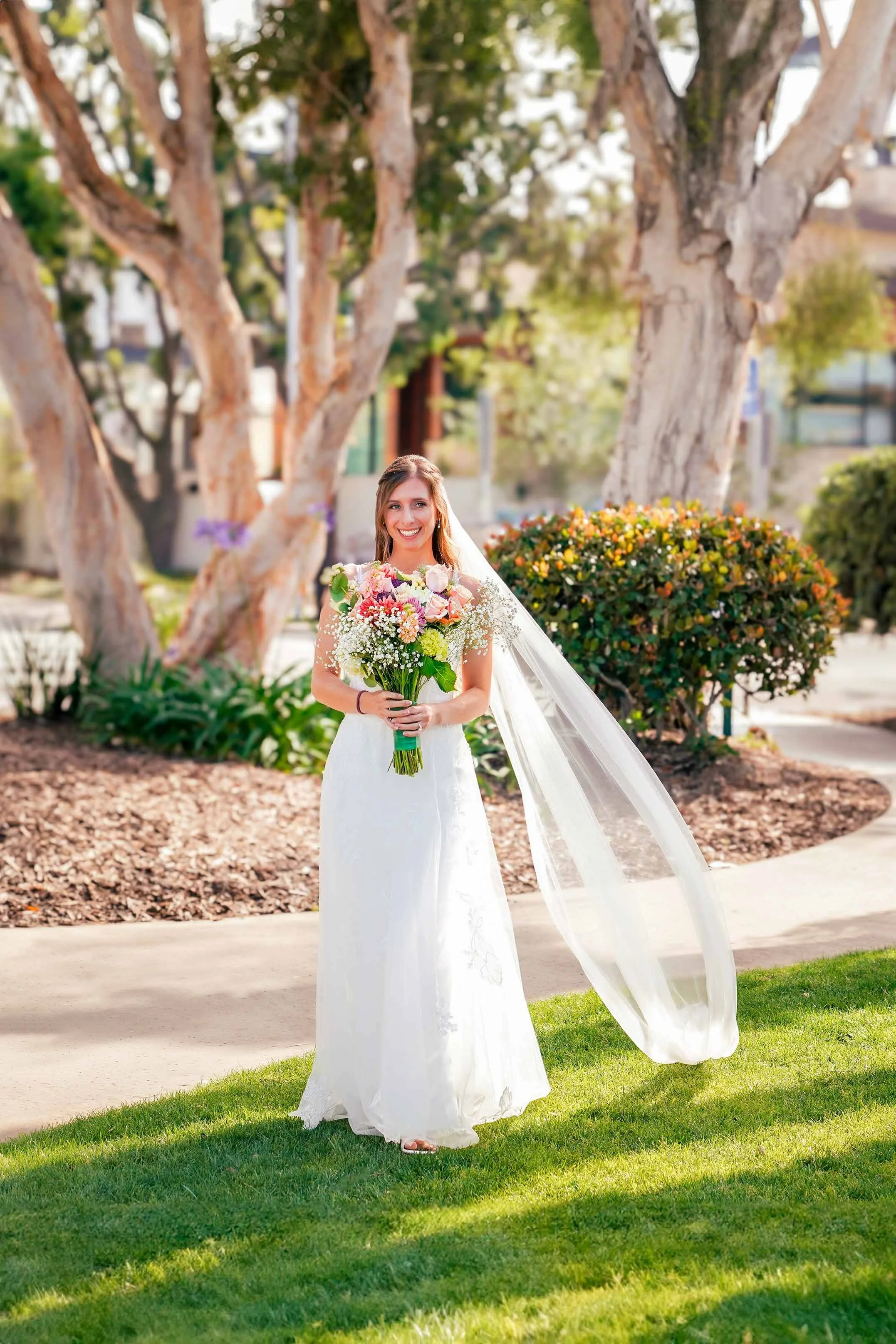 A smiling bride in a white lace wedding gown and a long, flowing veil stands outdoors on a sunny day in Coronado, holding a large, colorful floral bouquet.


