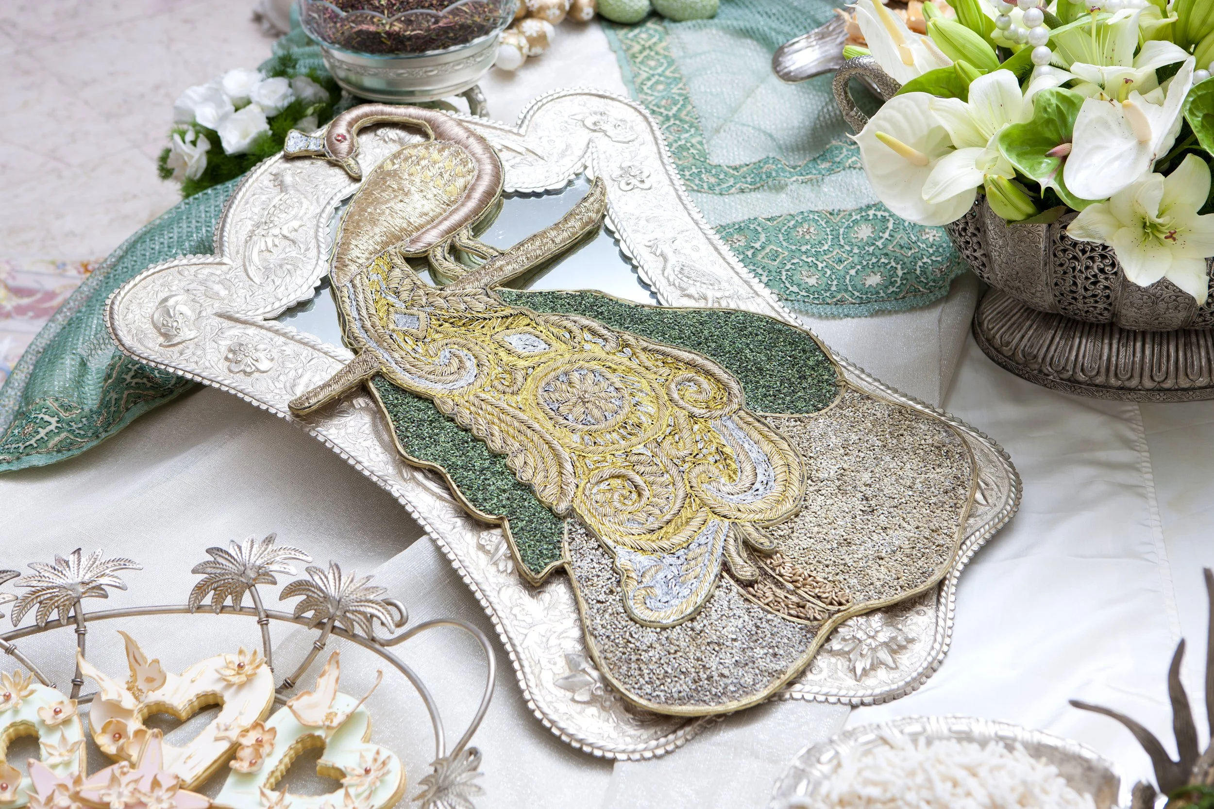 An ornate, gold and green embroidered peacock decoration with mirror accents, displayed on a silver platter as part of a formal table setting with lace linens and white flowers, likely for a Persian wedding ceremony Sofreh Aghd.