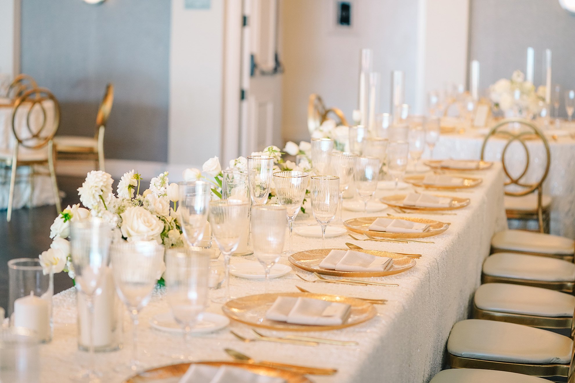 A long, formal dining table set for an event with white textured tablecloths, gold charger plates, white napkins, clear glassware, and white floral arrangements, with gold chiavari chairs on the sides.