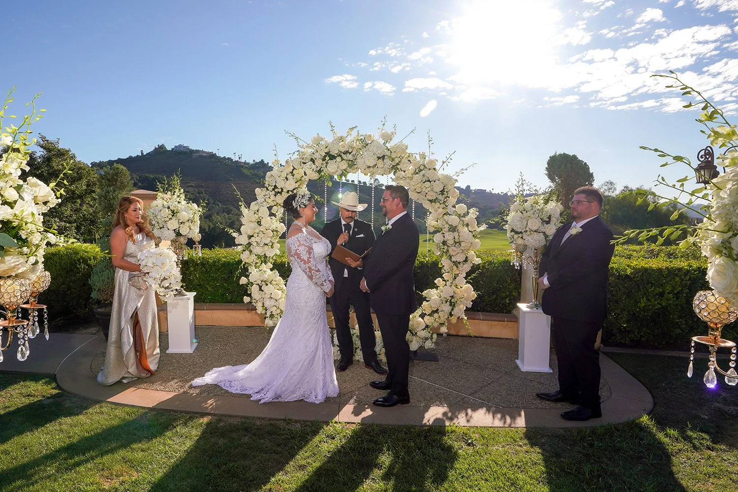 A bride and groom exchange vows during an outdoor wedding ceremony under a large, circular white floral arch. The bride, in a lace gown and sparkling headpiece, faces the groom, who is in a black tuxedo. Between them, an officiant in a cowboy hat rea