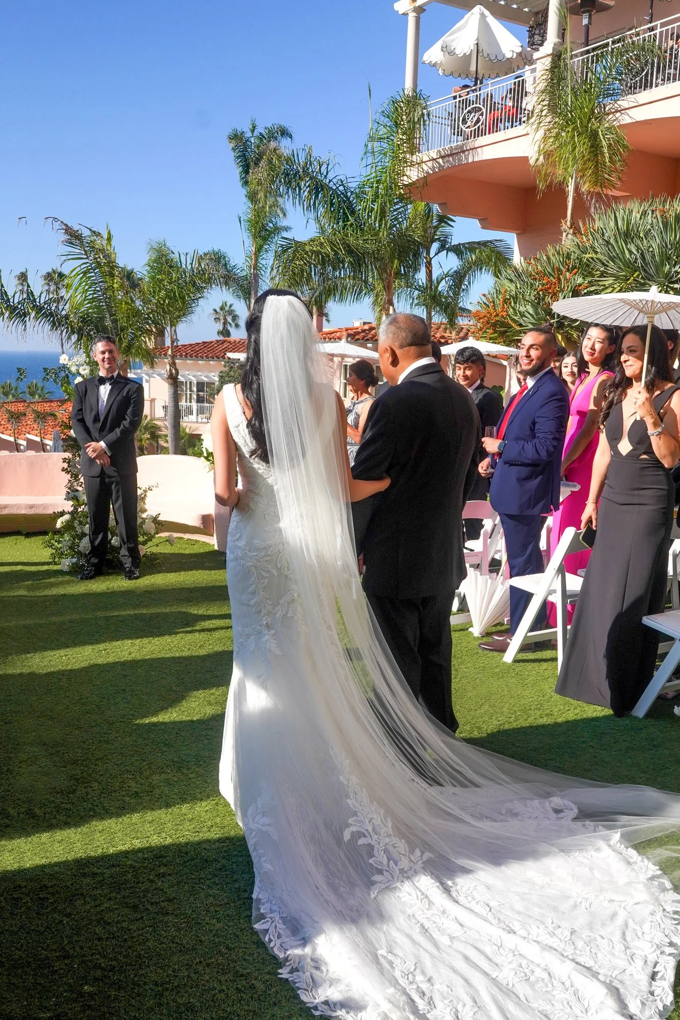 An outdoor photograph of a bride in a white lace wedding dress and long veil being escorted down a grassy aisle during a wedding ceremony. The bride is viewed from behind, walking alongside a man in a black suit. The groom stands to the left in a tux