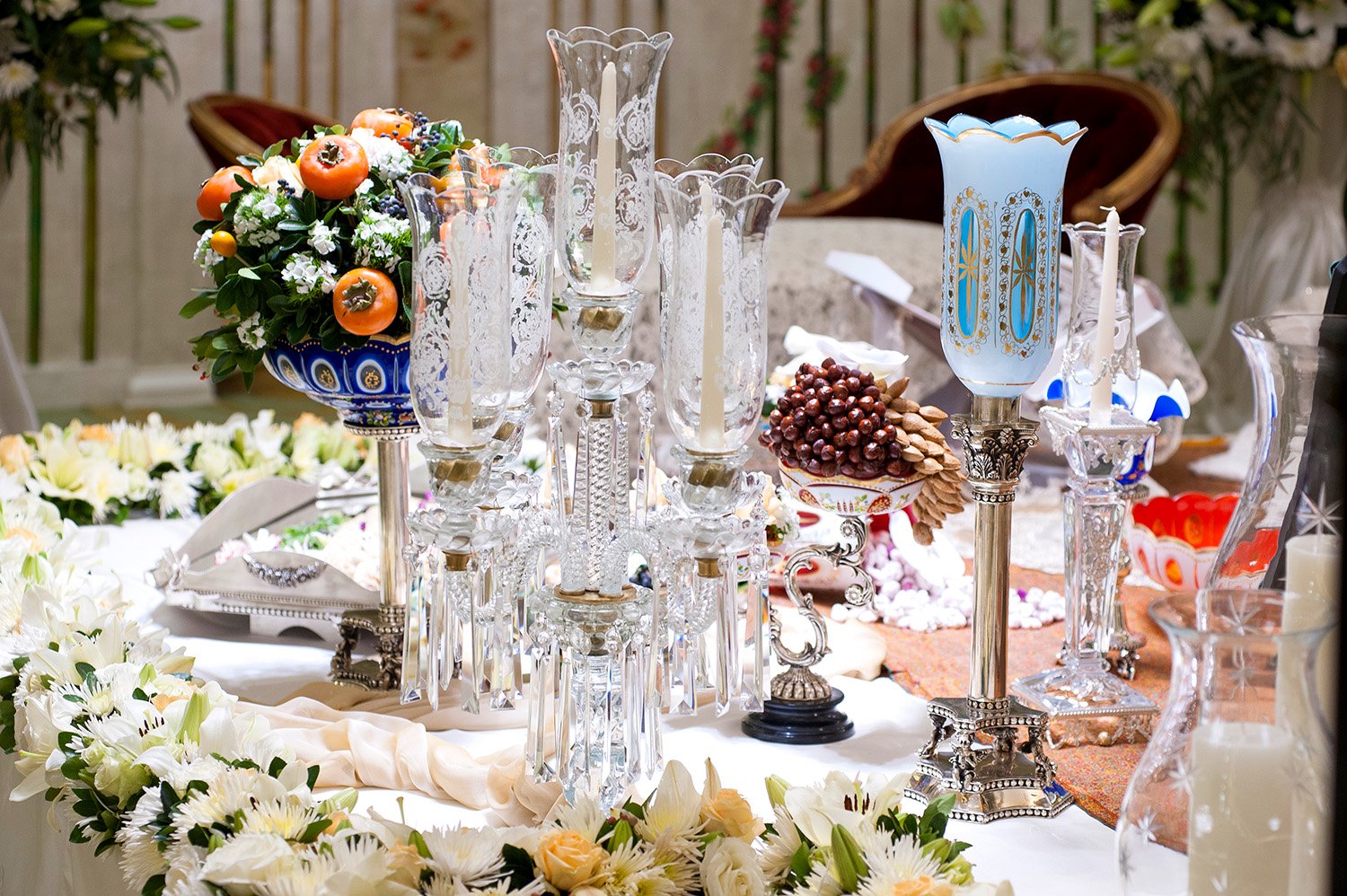 An ornate Persian wedding table setting (Sofreh Aghd) featuring crystal candelabras, a blue and gold candle holder, a bowl of nuts and dried fruits, and extensive white and orange floral arrangements.