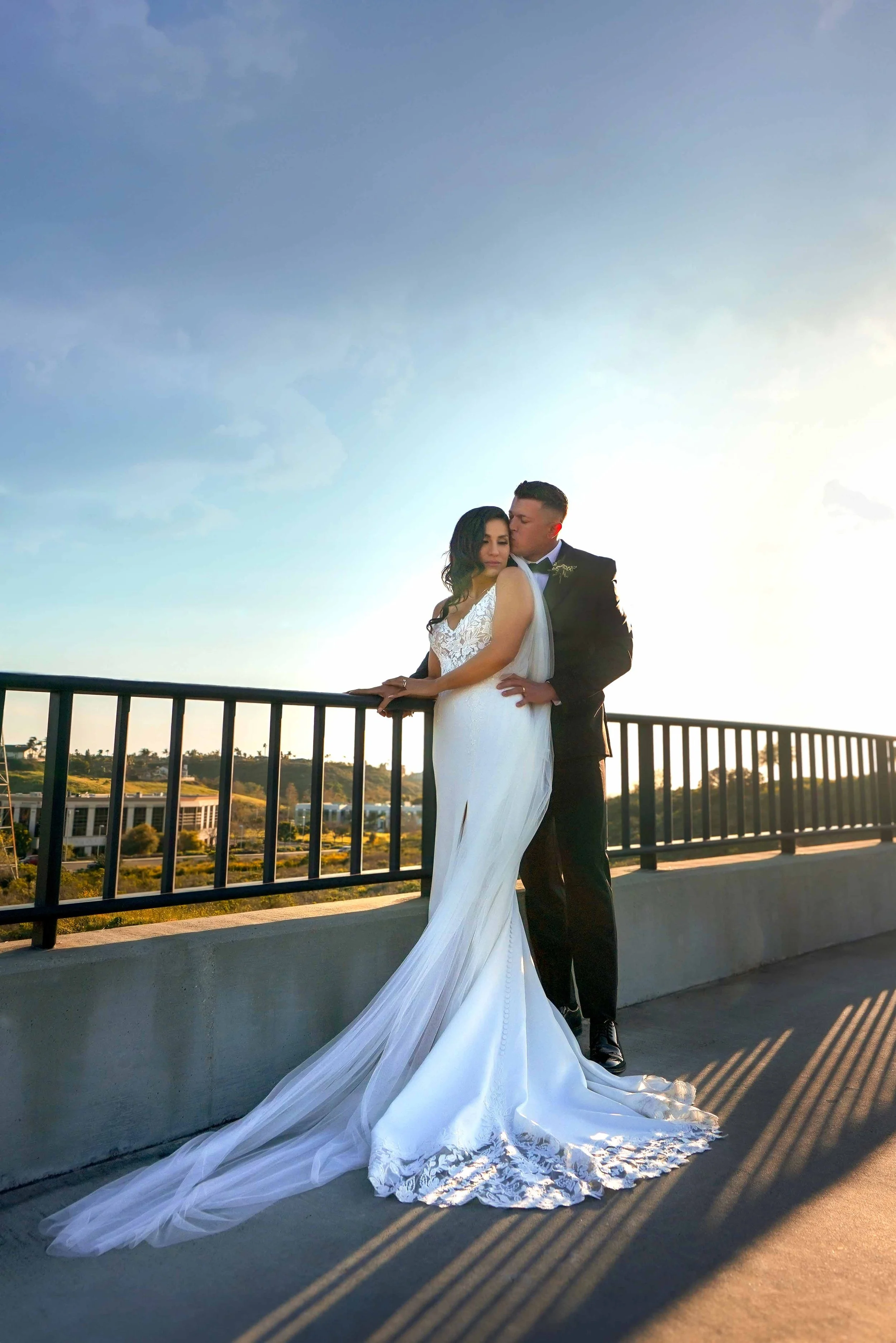 A full-length outdoor photograph of a newly married couple posing on an elevated walkway during sunset. The groom, in a dark suit or tuxedo, stands behind the bride and kisses her forehead. The bride wears a white lace wedding dress with a long train