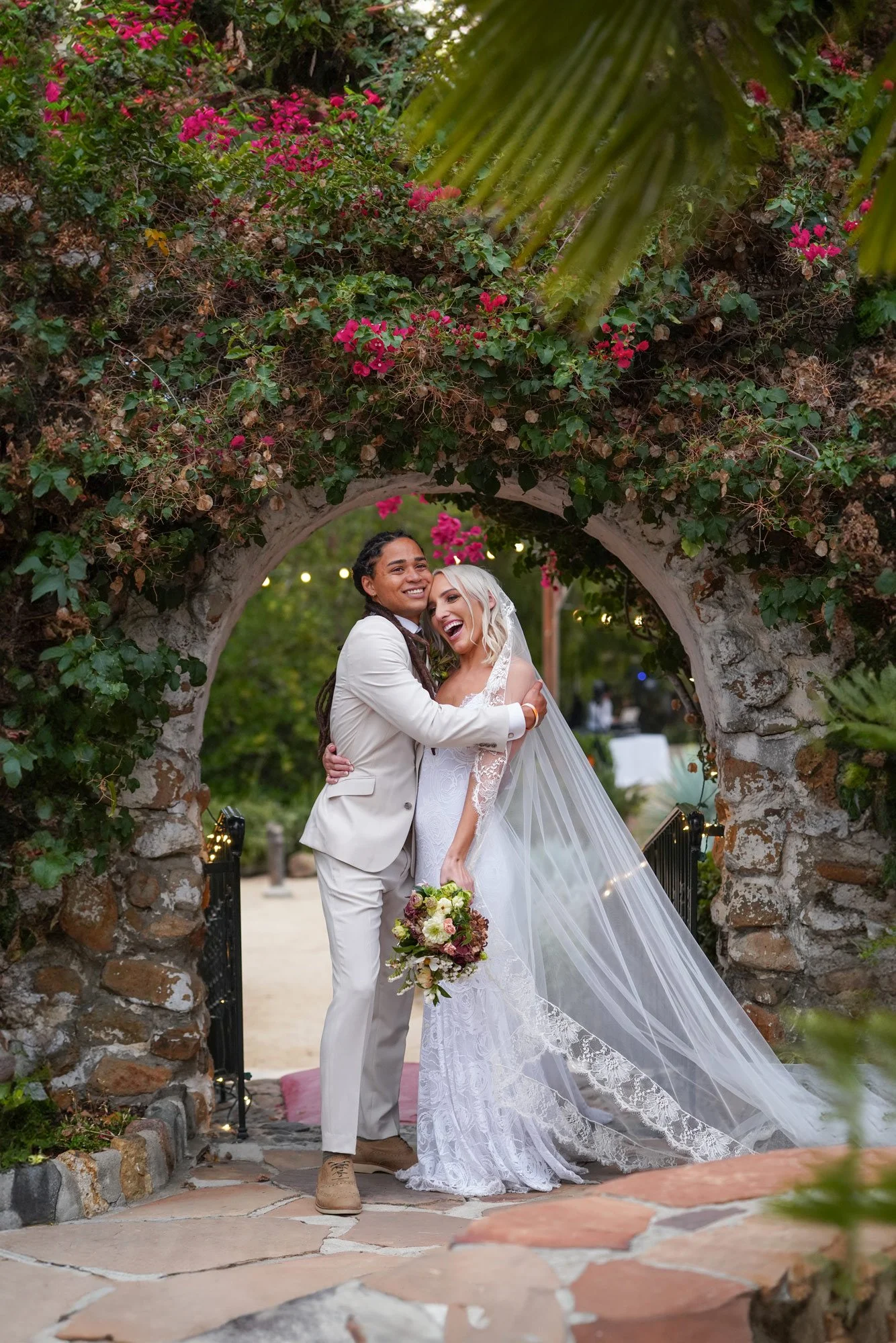 A photo of a newly married couple laughing and embracing under a stone archway covered in pink bougainvillea flowers, with the groom in a cream-colored suit and the bride in a white lace wedding dress with a long, lace-trimmed veil at Leo Garriloo, C