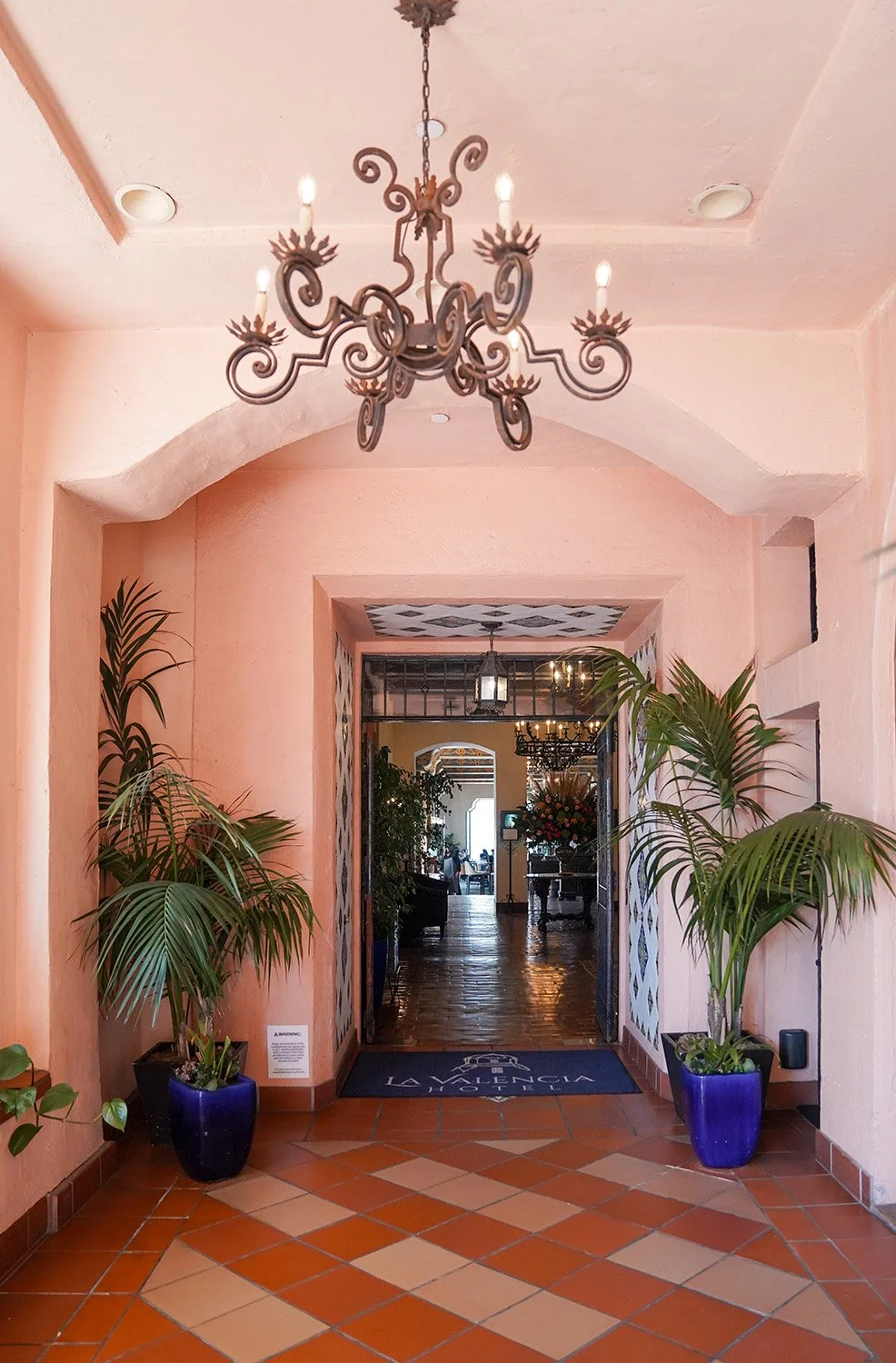 The entrance to the La Valencia Hotel features an arched doorway framed by pink stucco walls and blue potted palm plants, terracotta tile flooring, an ornate wrought iron chandelier, and a view into the hotel lobby.