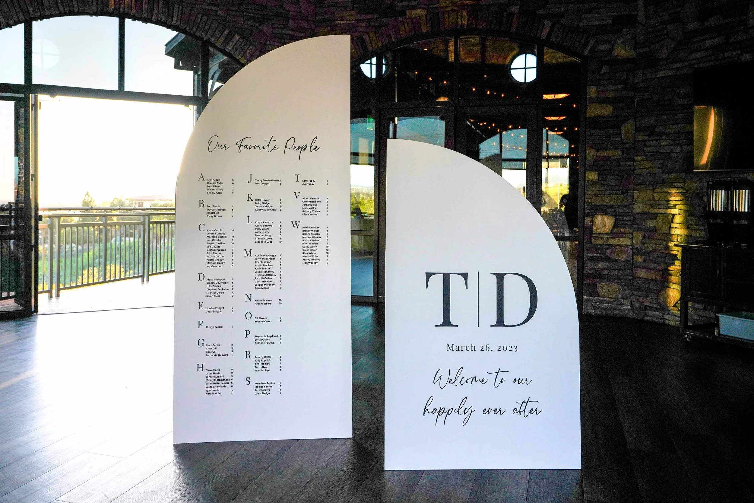 An indoor photograph of two large, arched white wedding signs set up on a dark wood floor. The left sign functions as a seating chart titled "Our Favorite People" with guest names listed alphabetically. The right sign displays the large black initial