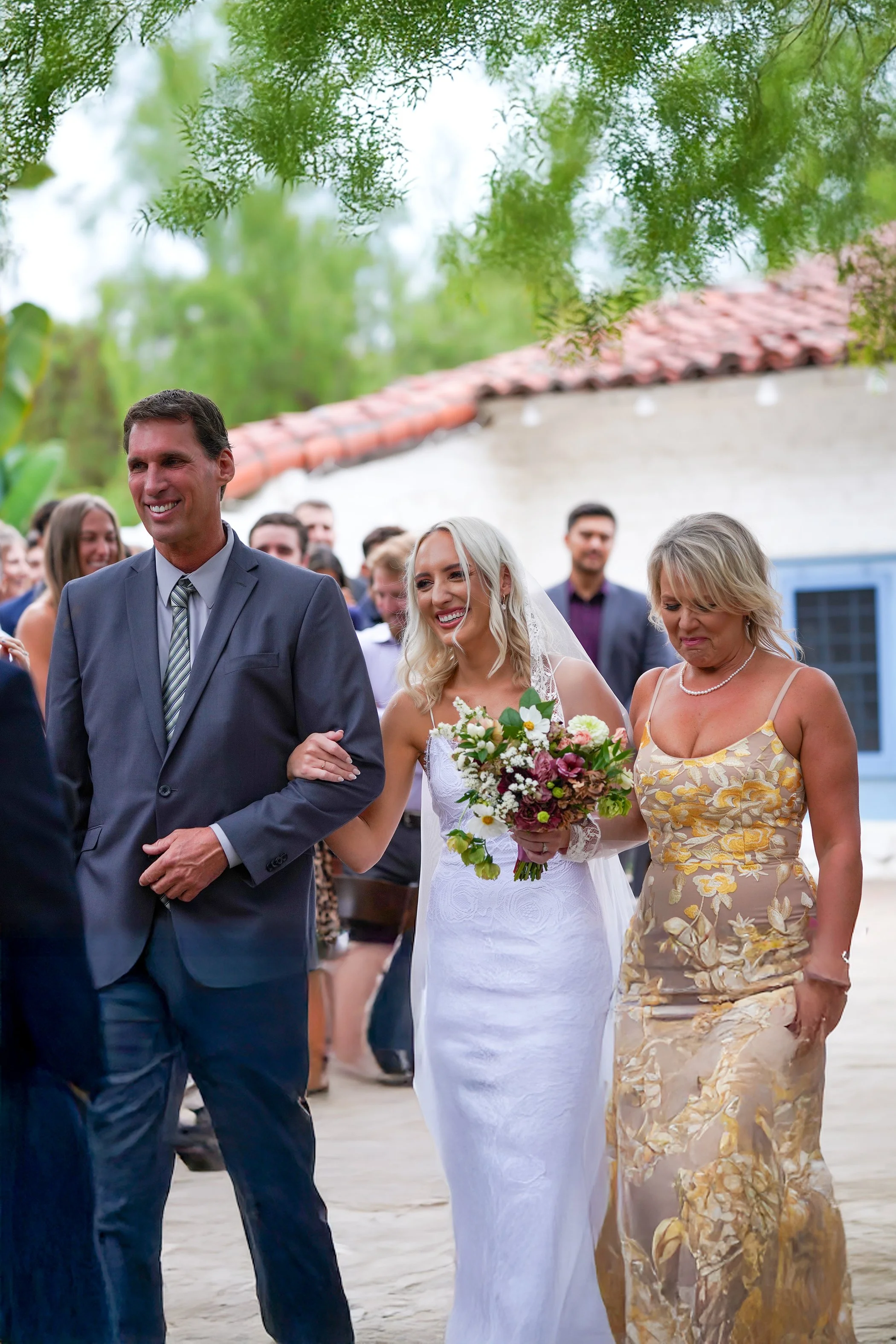 Bride in a white lace gown smiling while being walked down the aisle by her parents at an outdoor ceremony."
