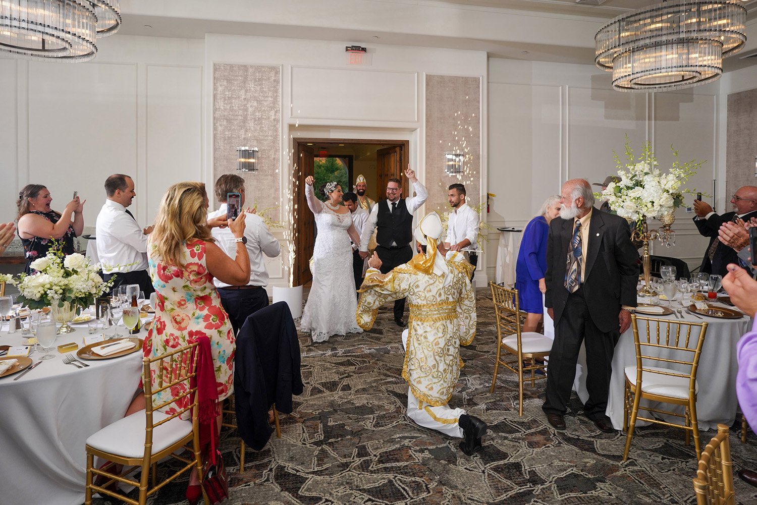 An indoor photograph capturing the enthusiastic entry of a newly married couple into their wedding reception at The Maderas Golf Club. The bride wears a white wedding dress, and the groom wears a black vest and white shirt. They enter through an arch