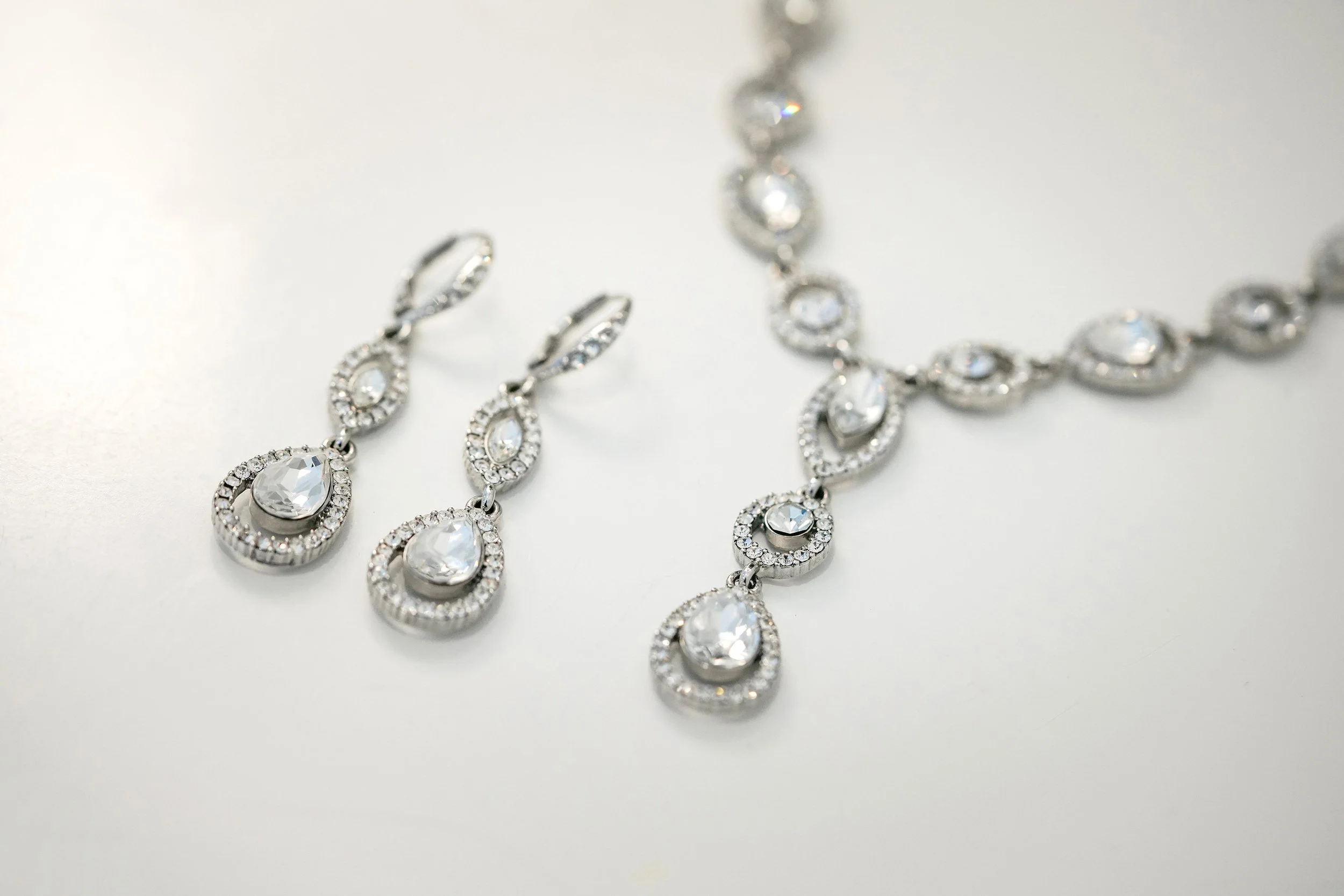 A set of silver-toned bridal jewelry including drop earrings and a necklace, featuring pear-shaped and oval clear stones encircled by smaller pave-set gems, all arranged on a white surface.


