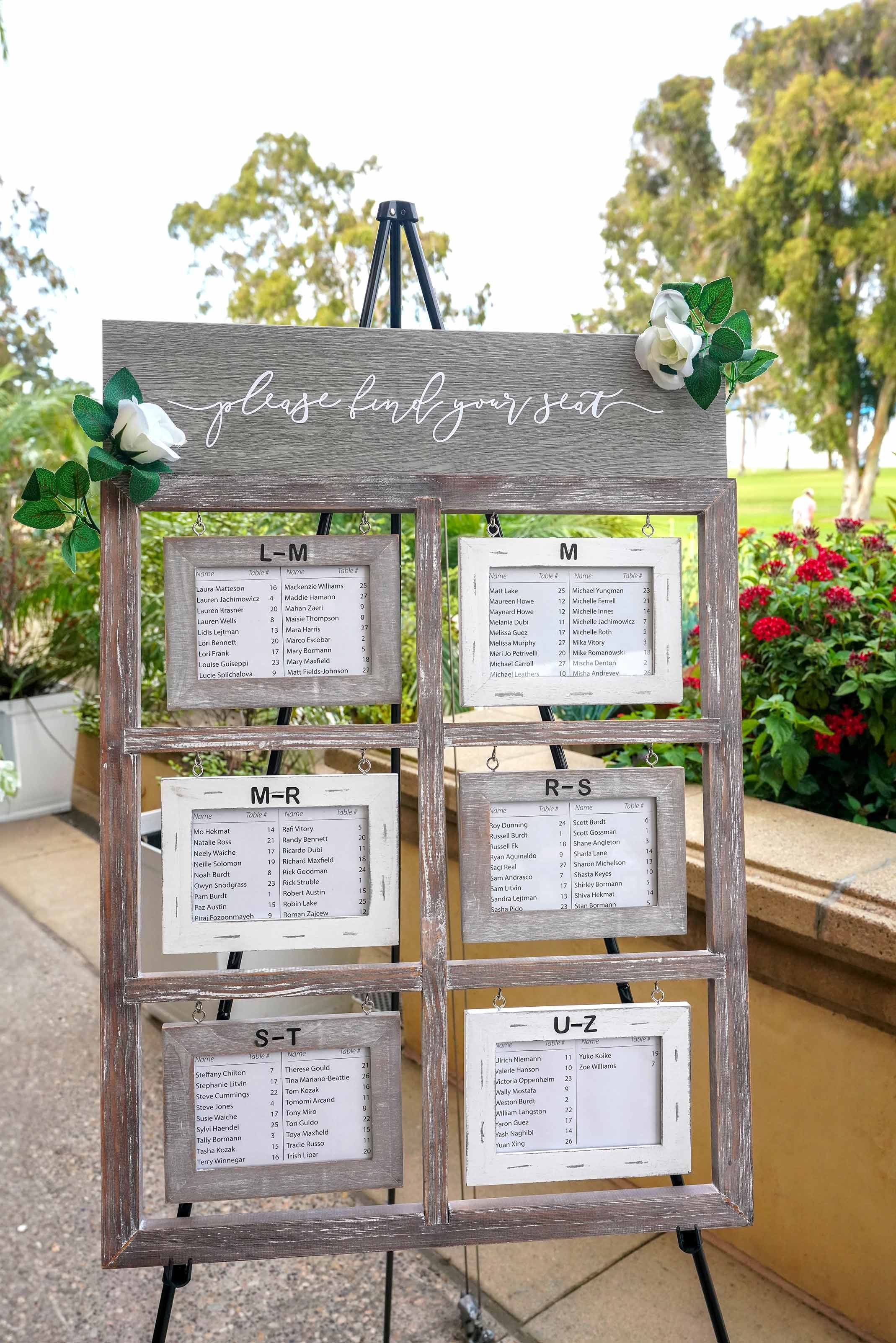 A rustic, gray wooden wedding seating chart display designed to look like a multi-pane window, with white script text at the top reading "please find your seat" and framed lists of guest names organized alphabetically by last name range (L-M through 