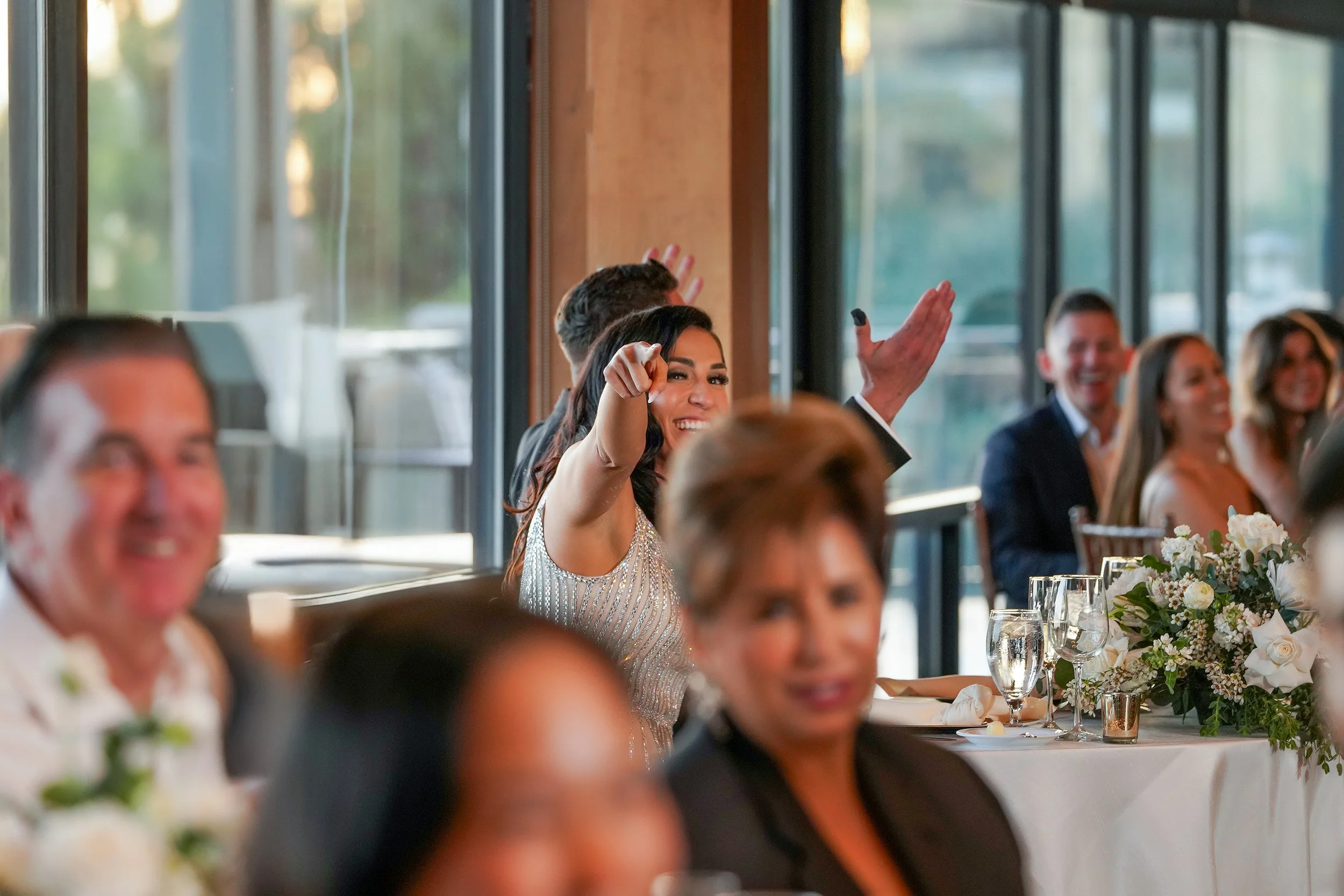 Candid bride reaction at The Crossings wedding reception in Carlsbad, featuring elegant indoor decor and white floral centerpieces. Bride reacting with a smile and pointing during an indoor wedding reception with guests seated at tables.