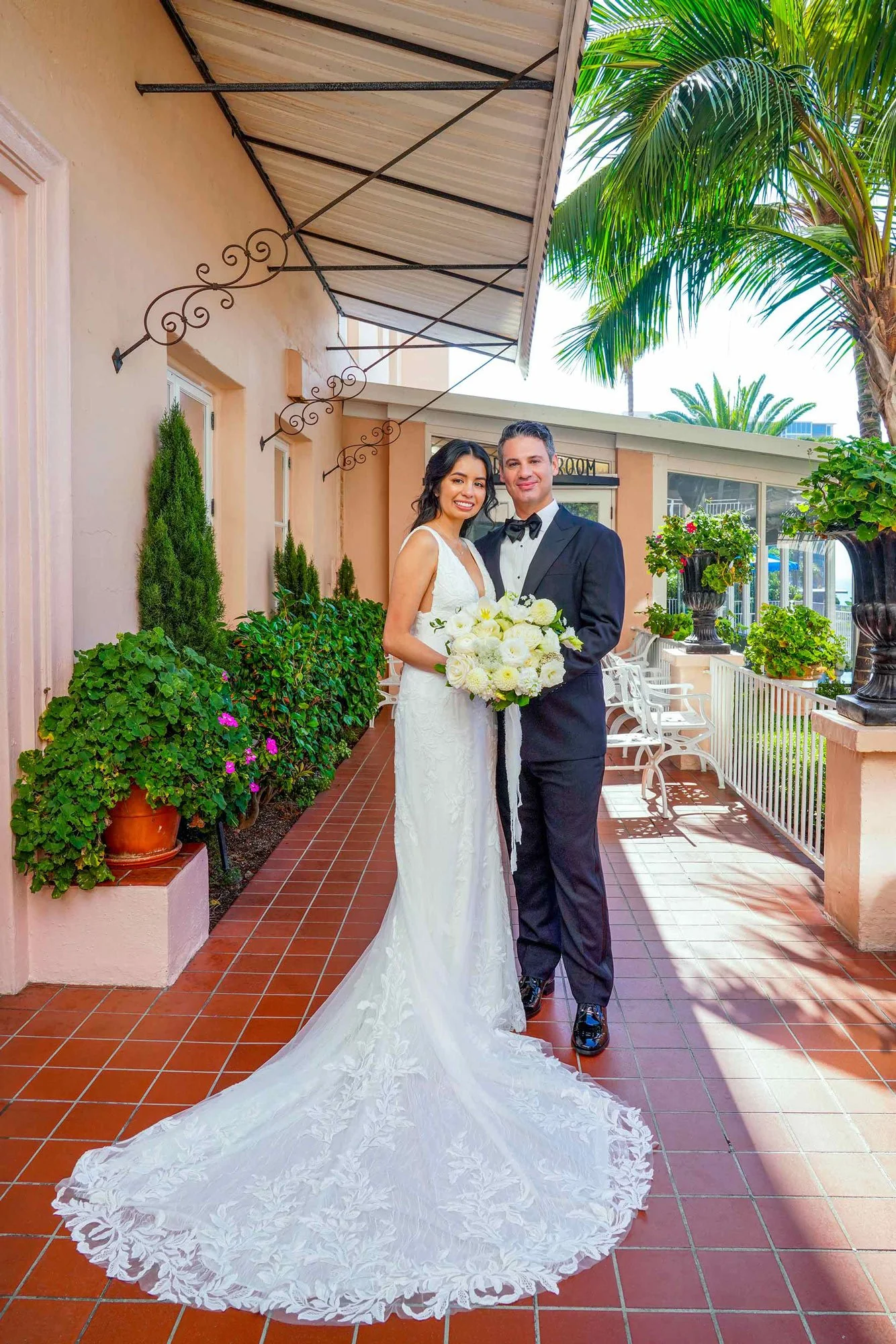 A full-length outdoor photograph of a newly married couple posing together. They are standing on a terracotta-tiled patio in front of a pink building at La Valencia Hotel with an awning, surrounded by green plants, including tall palm trees and white