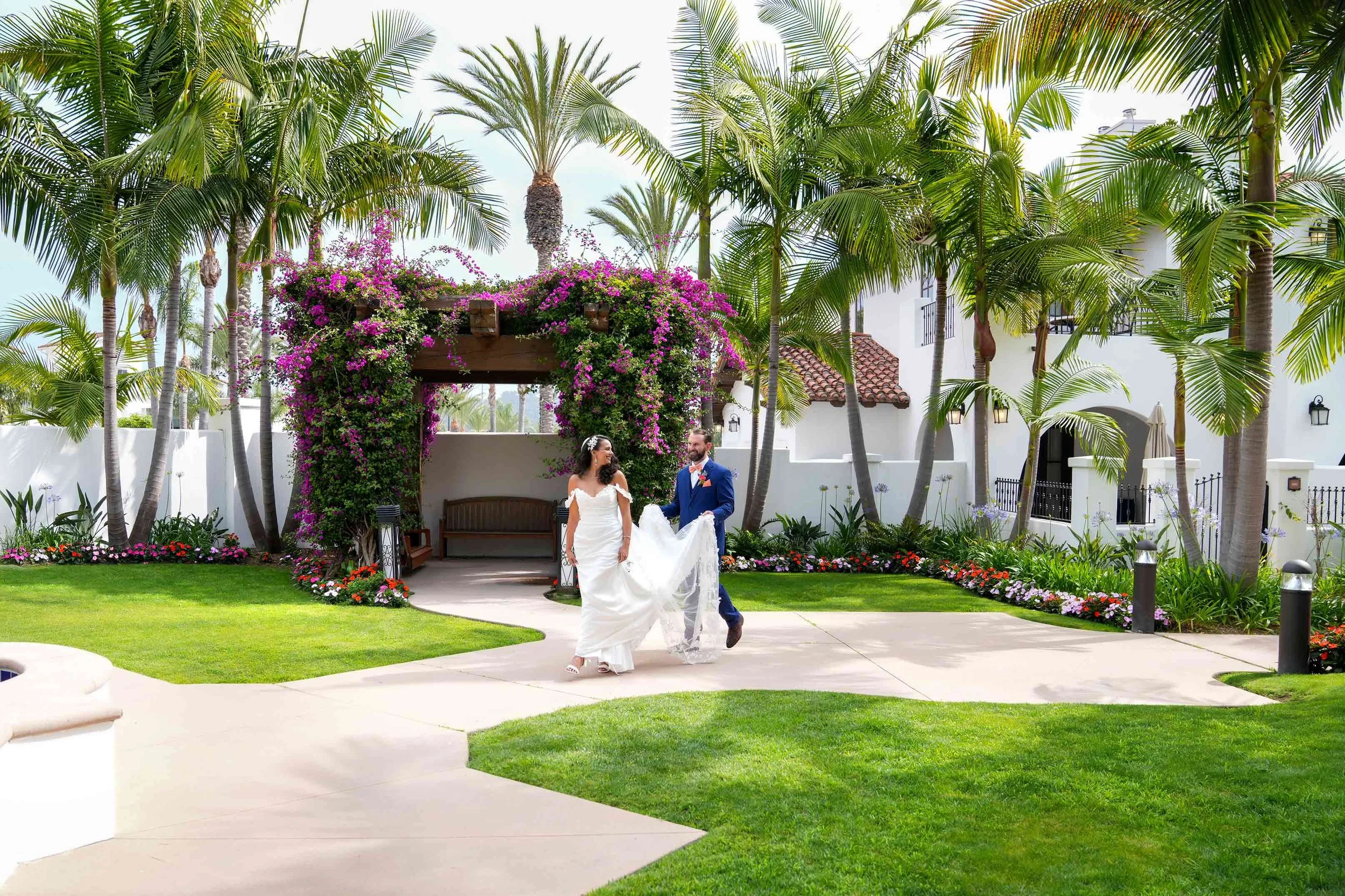 A bride in a white gown and a groom in a blue suit walk along a paved path in a lush garden setting surrounded by palm trees and white buildings in Omni La Costa, passing under a wooden arch covered in pink flowers.