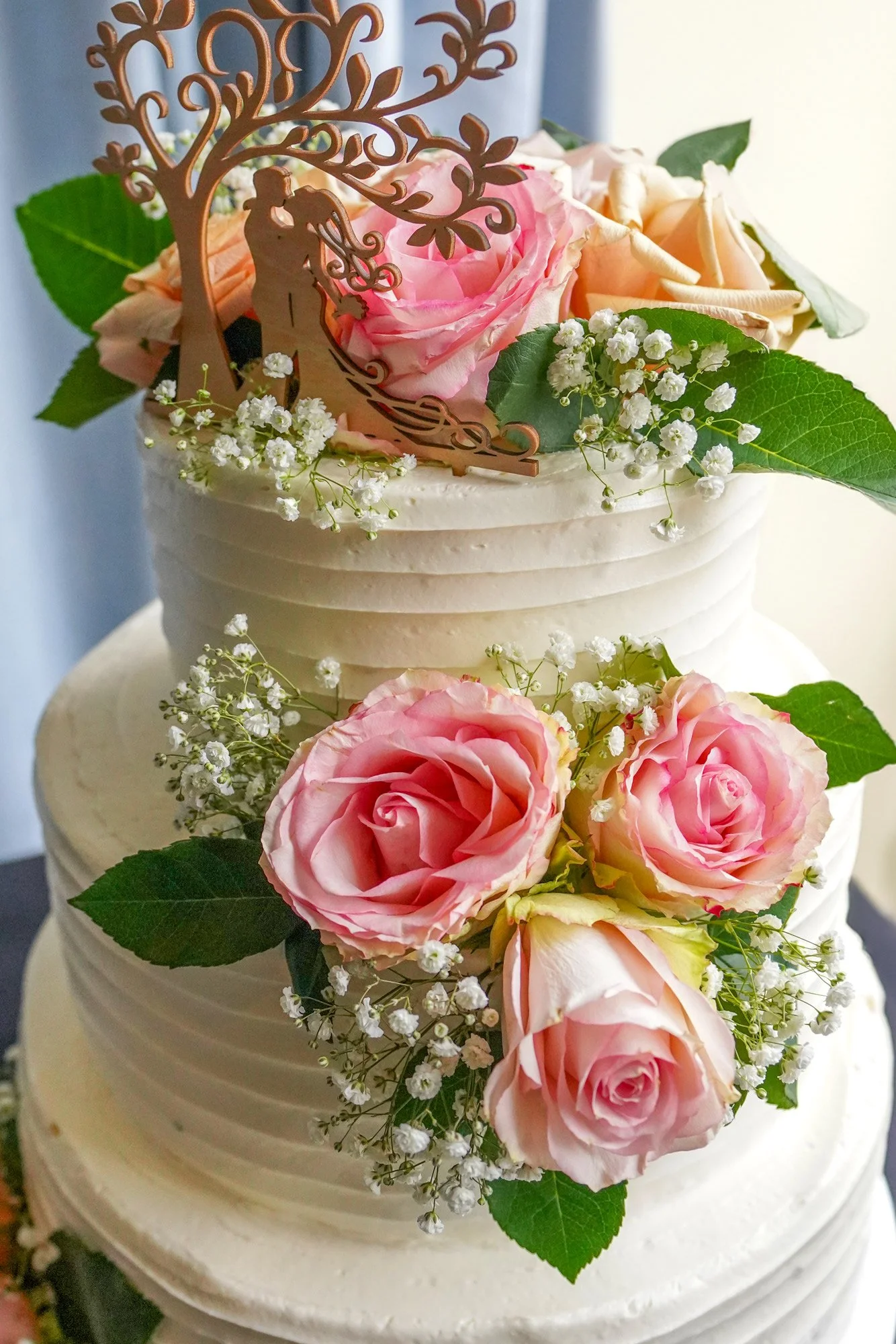 A close-up of a two-tiered white wedding cake with horizontal textured frosting, decorated with fresh pink and peach roses, white baby's breath, and a wooden cake topper of a couple kissing under a tree.



