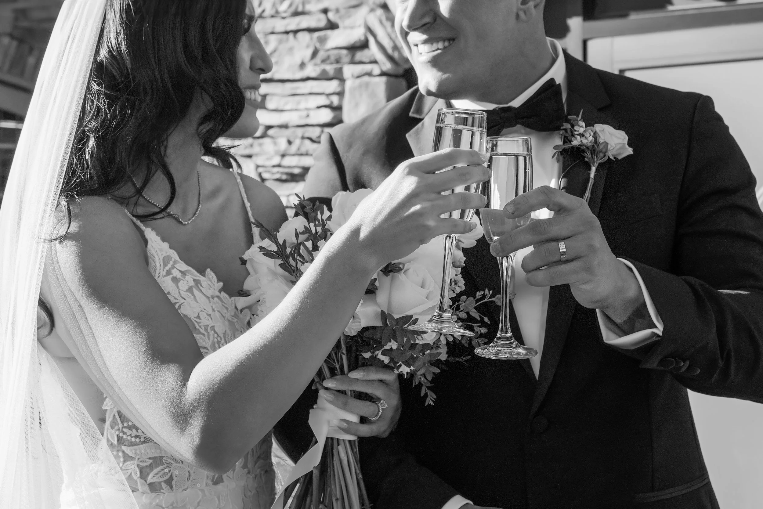 A bride in a lace gown and a groom in a black tuxedo share a celebratory champagne toast, featuring a close-up of their wedding rings and a white rose bouquet.