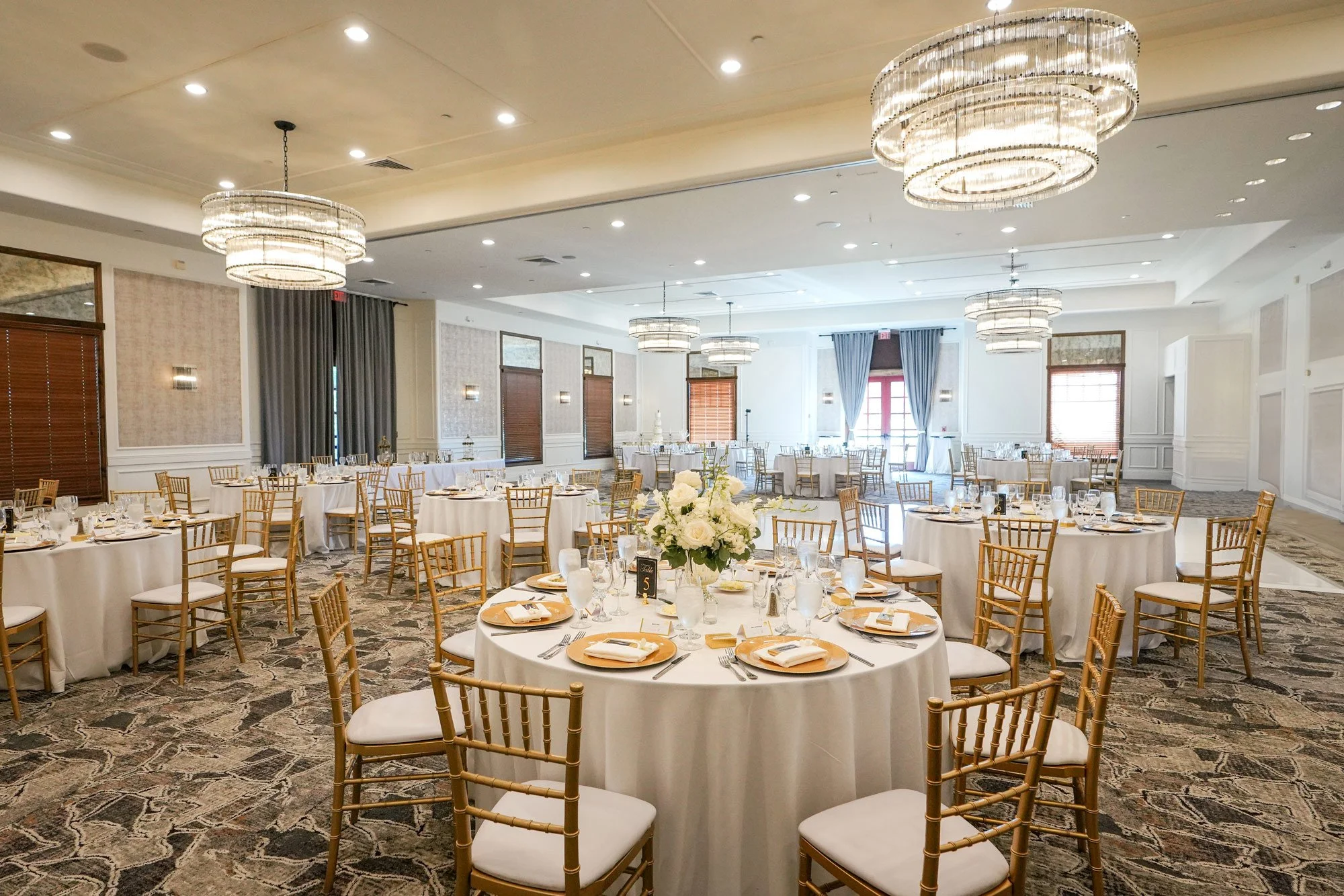 An indoor photograph of an elegant wedding reception setup in a banquet hall. Round tables are set with white linens, gold charger plates, and white floral centerpieces. Gold Chiavari chairs surround the tables, and large, modern crystal chandeliers 