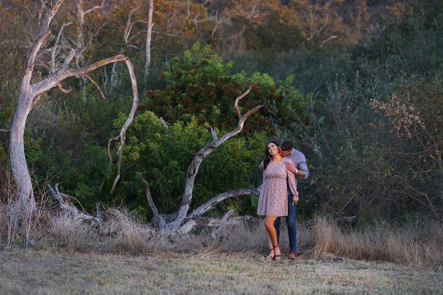 An outdoor photograph of a couple embracing in a natural, overgrown area during warm evening light. The woman wears a short, light-colored, floral dress, and the man, in a grey shirt and blue jeans, stands behind her, appearing to kiss her on the nec