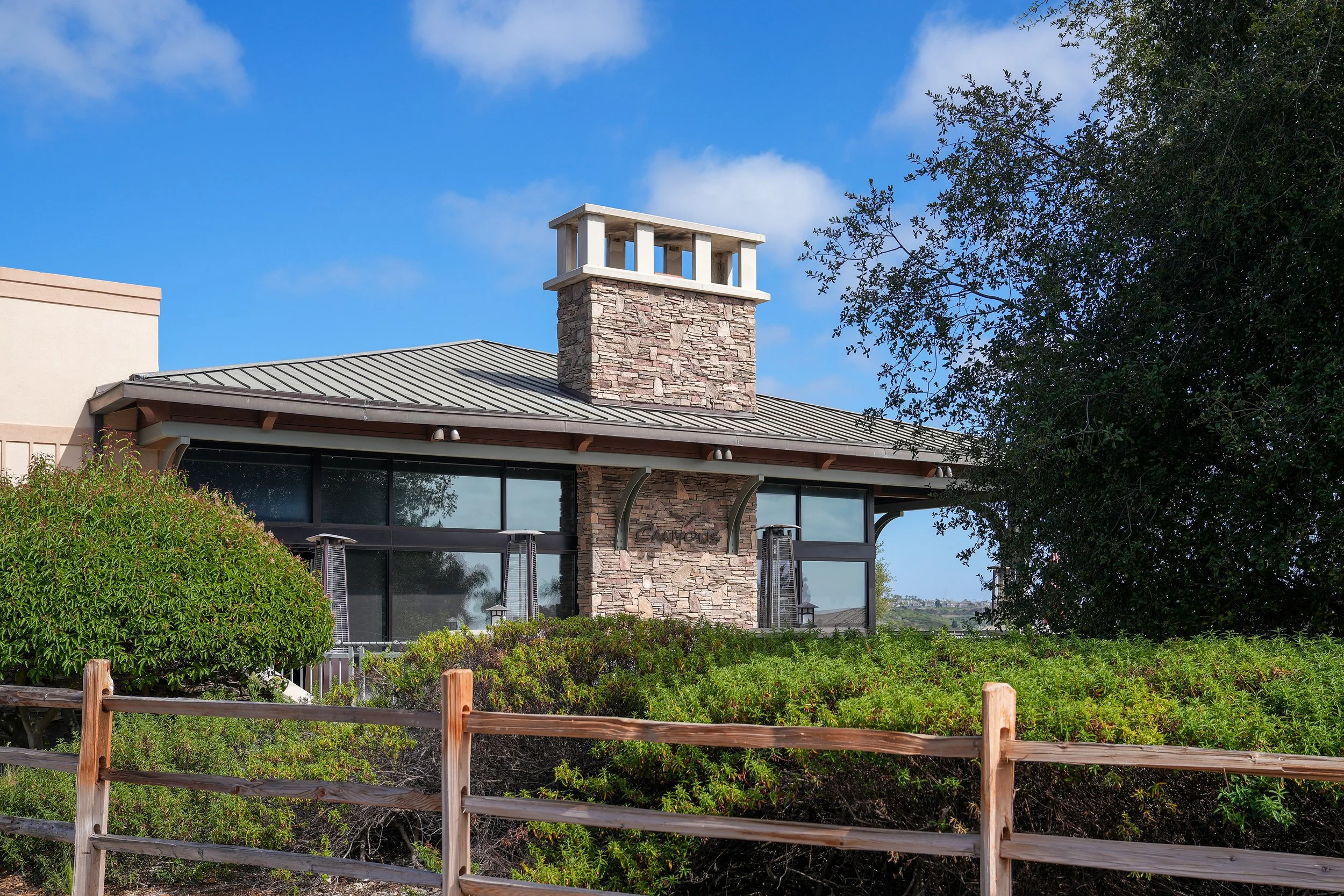The exterior of the Canyons Restaurant clubhouse at The Crossings at Carlsbad, a Craftsman-style stone and stucco building with a metal roof and a large chimney, surrounded by green hedges and trees under a blue sky.