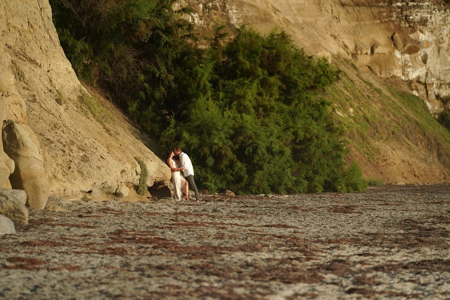 A couple embracing and kissing on a sandy beach below large, steep, sandy cliffs. The man wears a white long-sleeved shirt and dark pants, and the woman wears a light-colored dress. The cliffs are covered with dark green vegetation, and the foregroun