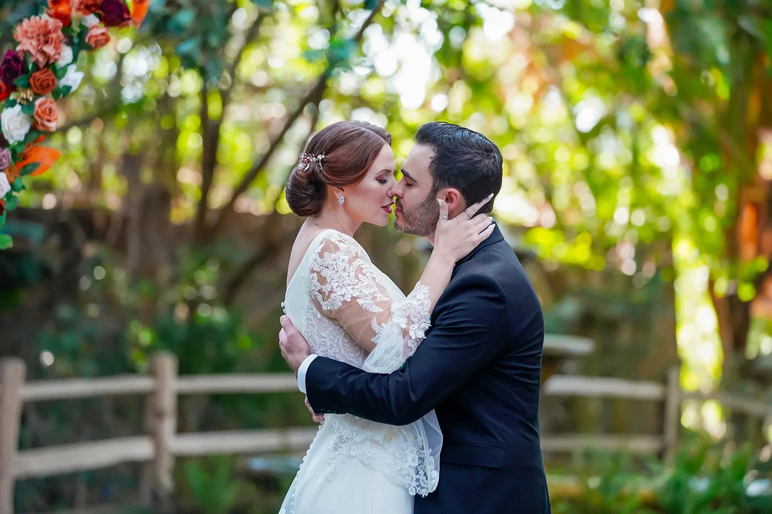 A bride and groom share an intimate moment outdoors, leaning in for a kiss. The bride, with her hair in a low bun and wearing an intricate lace-sleeved wedding gown, has her hand gently resting on the groom's neck. He is dressed in a classic black su