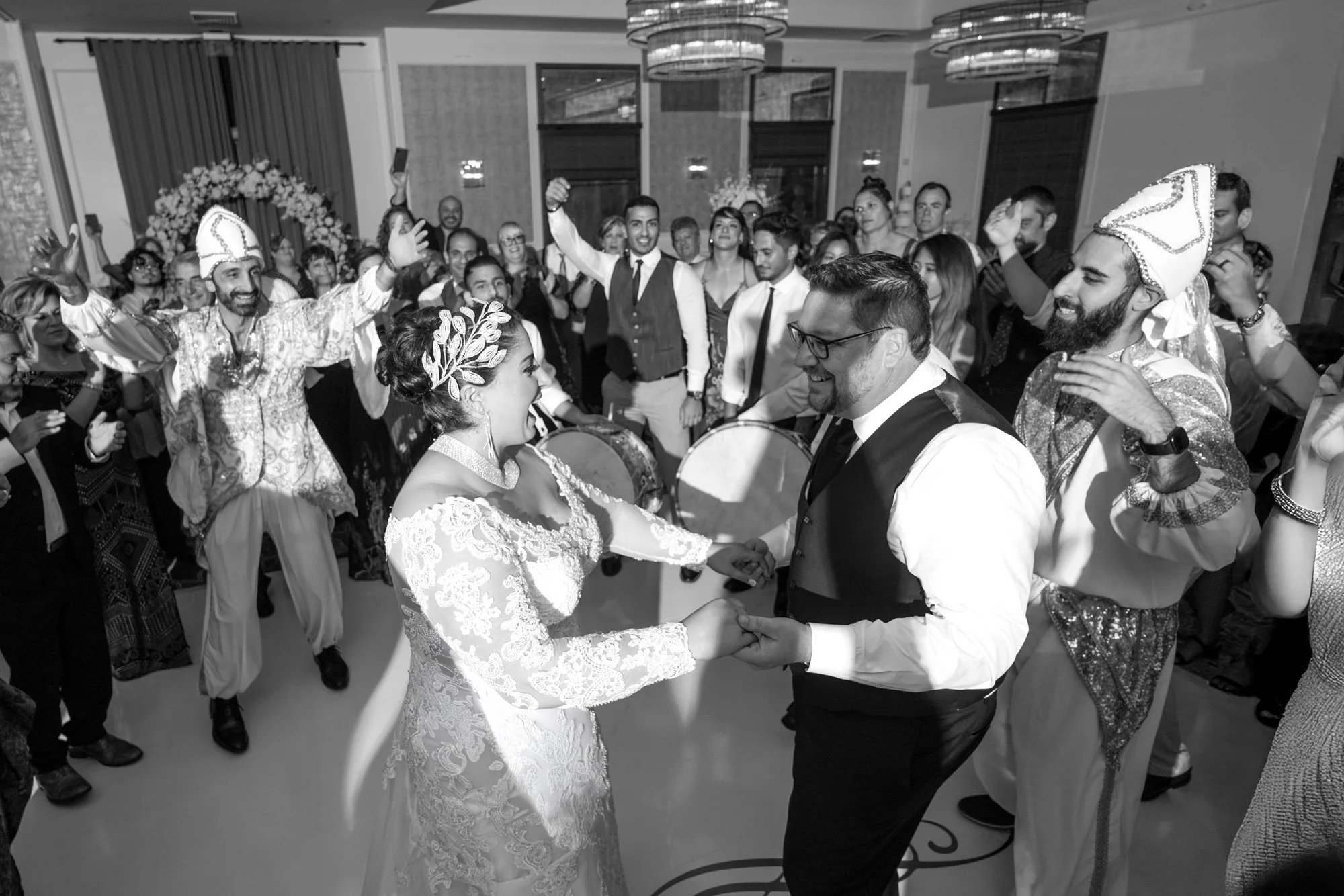 A black and white indoor photograph capturing a lively wedding reception celebration. The bride, in a white lace dress, and groom, in a black vest, are dancing and holding hands in the center of the image, smiling at each other. Guests surround them,