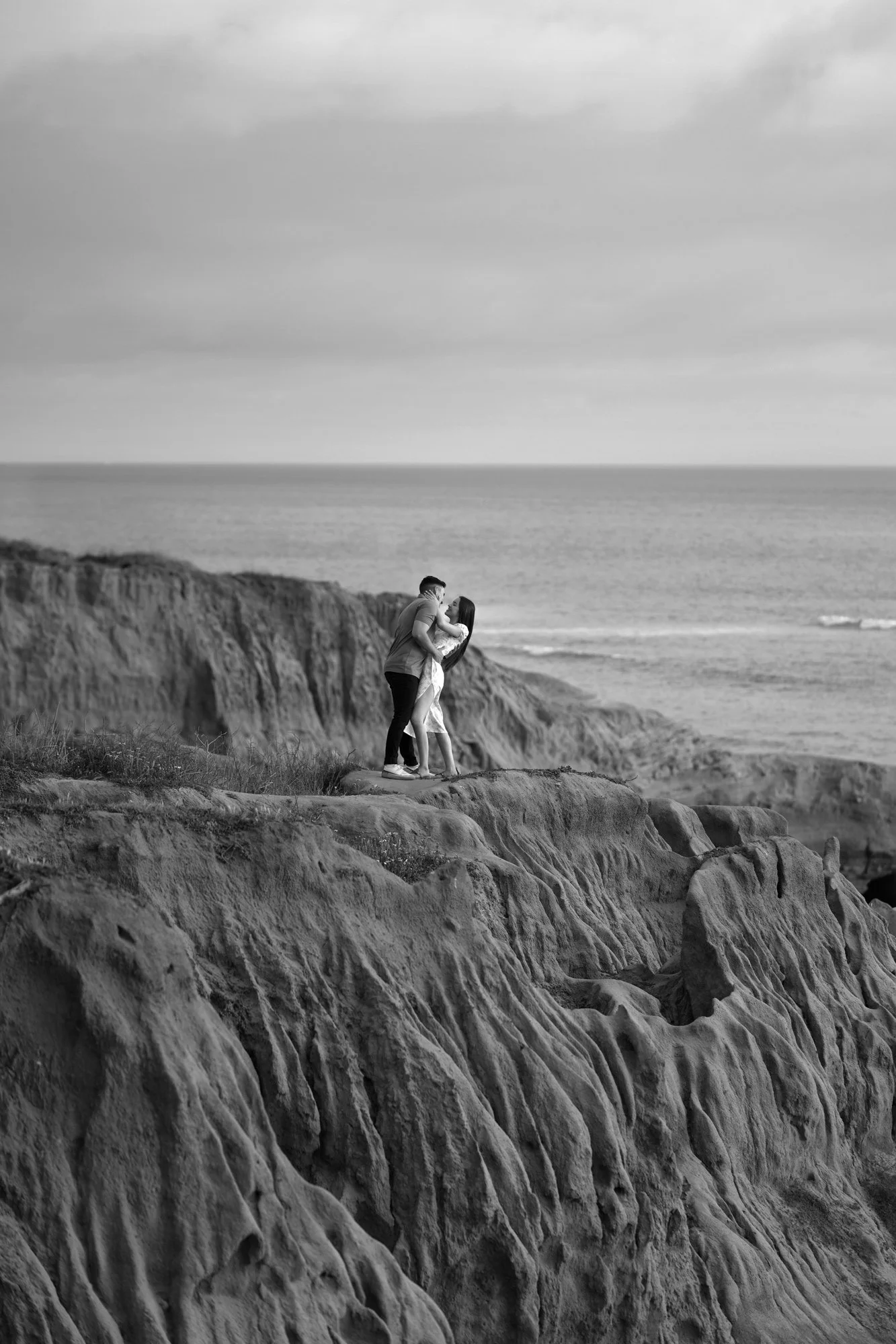 A black and white photograph of a couple embracing and kissing on a rugged, rocky cliff overlooking the ocean. The man wears a light shirt and dark pants, and the woman wears a light-colored dress. The sky is overcast.


