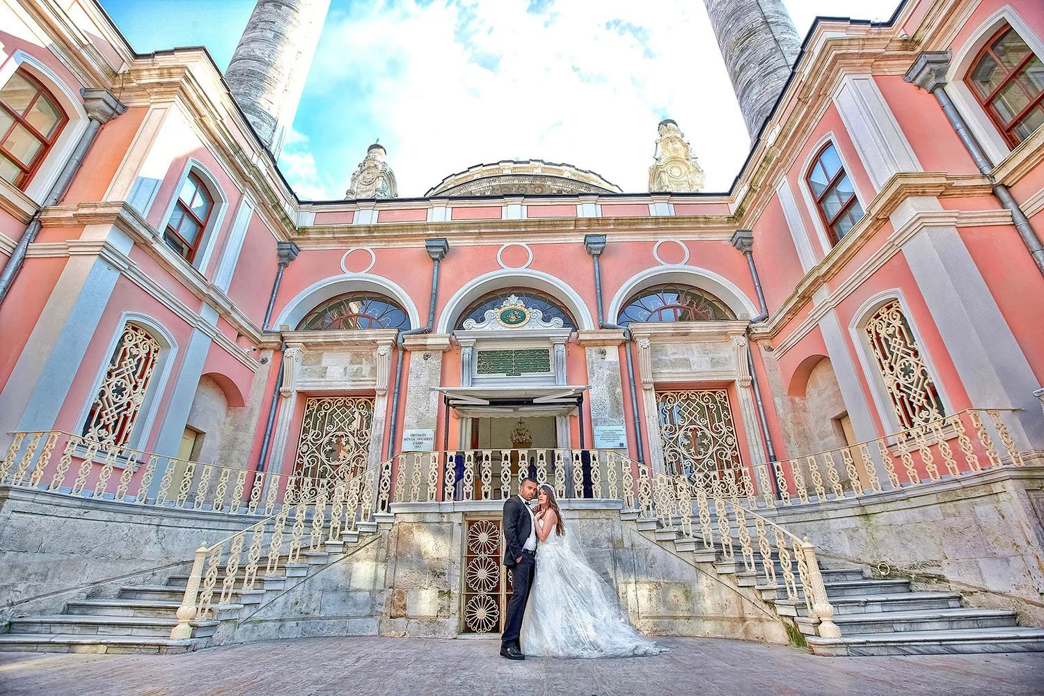 A bride and groom share a moment in front of the ornate Ortaköy Mosque in Istanbul, Turkey. The bride, in a voluminous white lace wedding gown with a long train, and the groom, in a dark suit, are centered at the base of the mosque's entrance steps. 