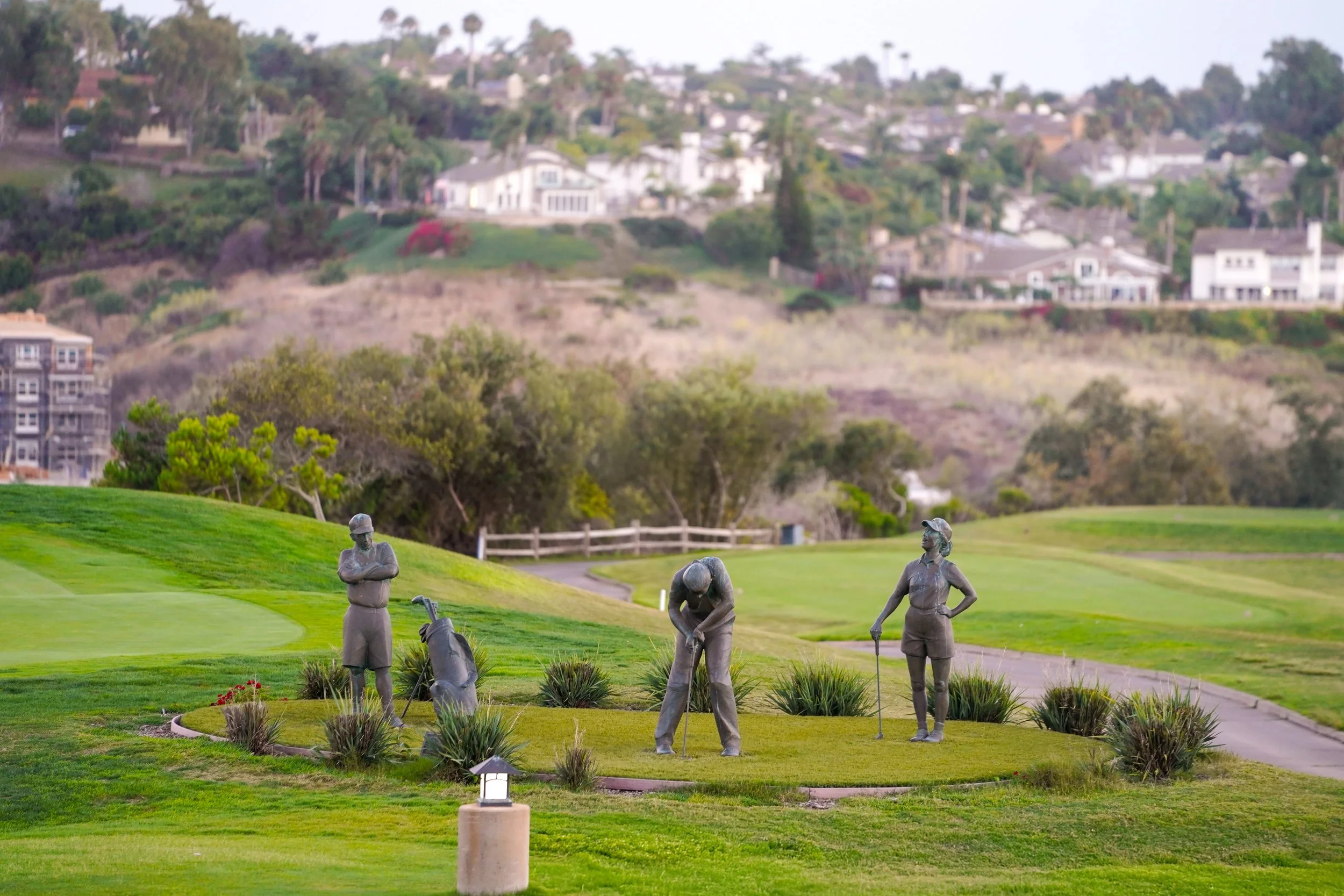 An outdoor display of three life-sized bronze golf statues (a man with crossed arms, a man bending to putt, and a woman with her hand on her hip) positioned in a garden bed on a green golf course, overlooking residential hills in the background. This