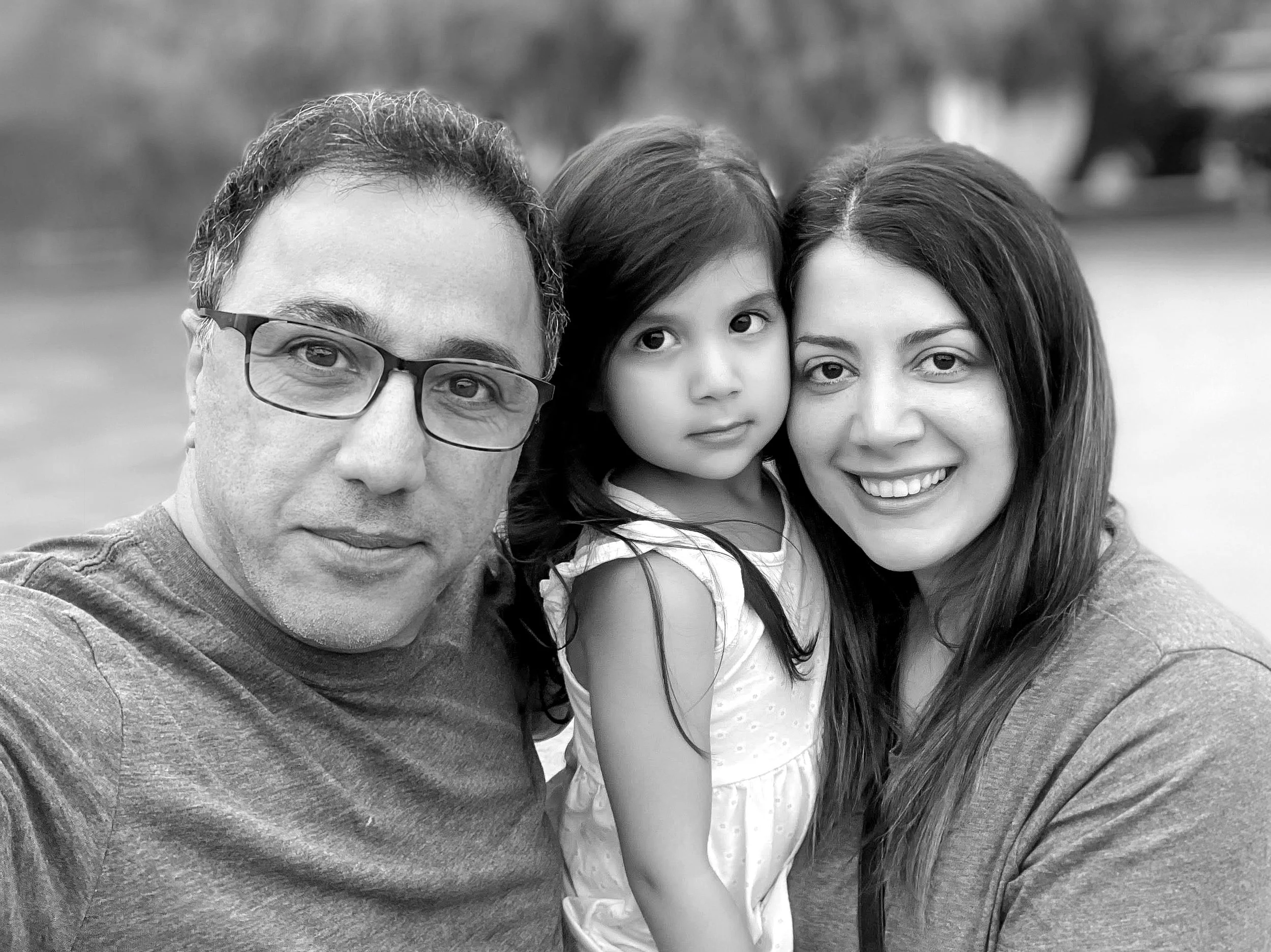 A black and white portrait photo of a family of three: a man with glasses, a young girl, and a woman. They are positioned closely together and looking towards the camera.