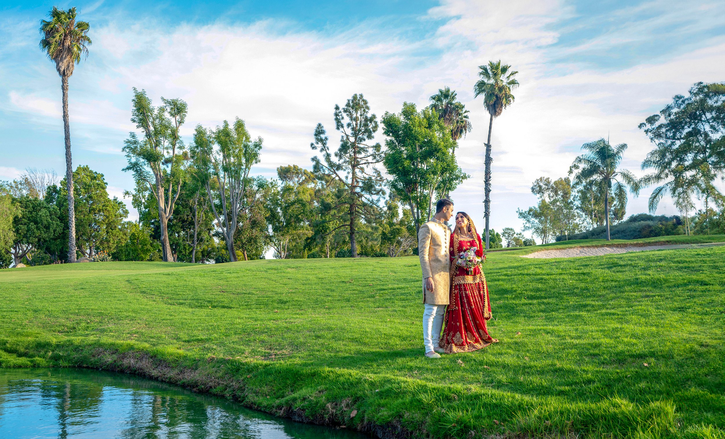 A bride in a red and gold lehenga and groom in a gold sherwani stand on a grassy hill beside a pond with palm trees in the background.