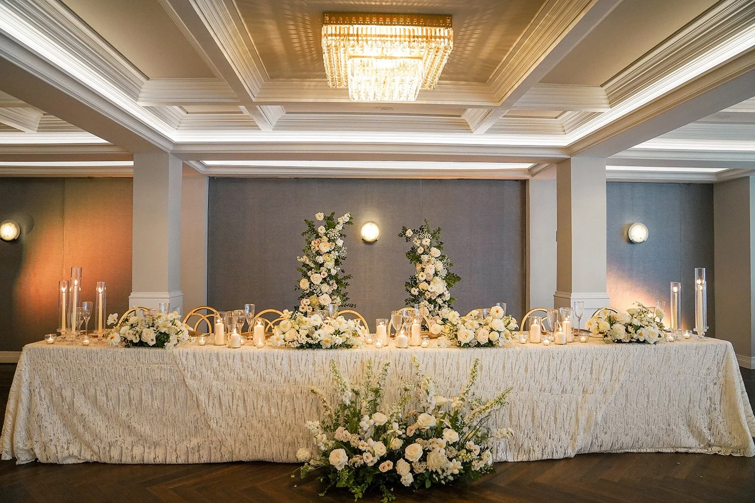 The venue features a refined interior with a coffered ceiling and a large, multi-tiered crystal chandelier providing warm, overhead lighting. The overall color palette is a classic mix of white, ivory, and gold, with muted grey wall panels, creating 