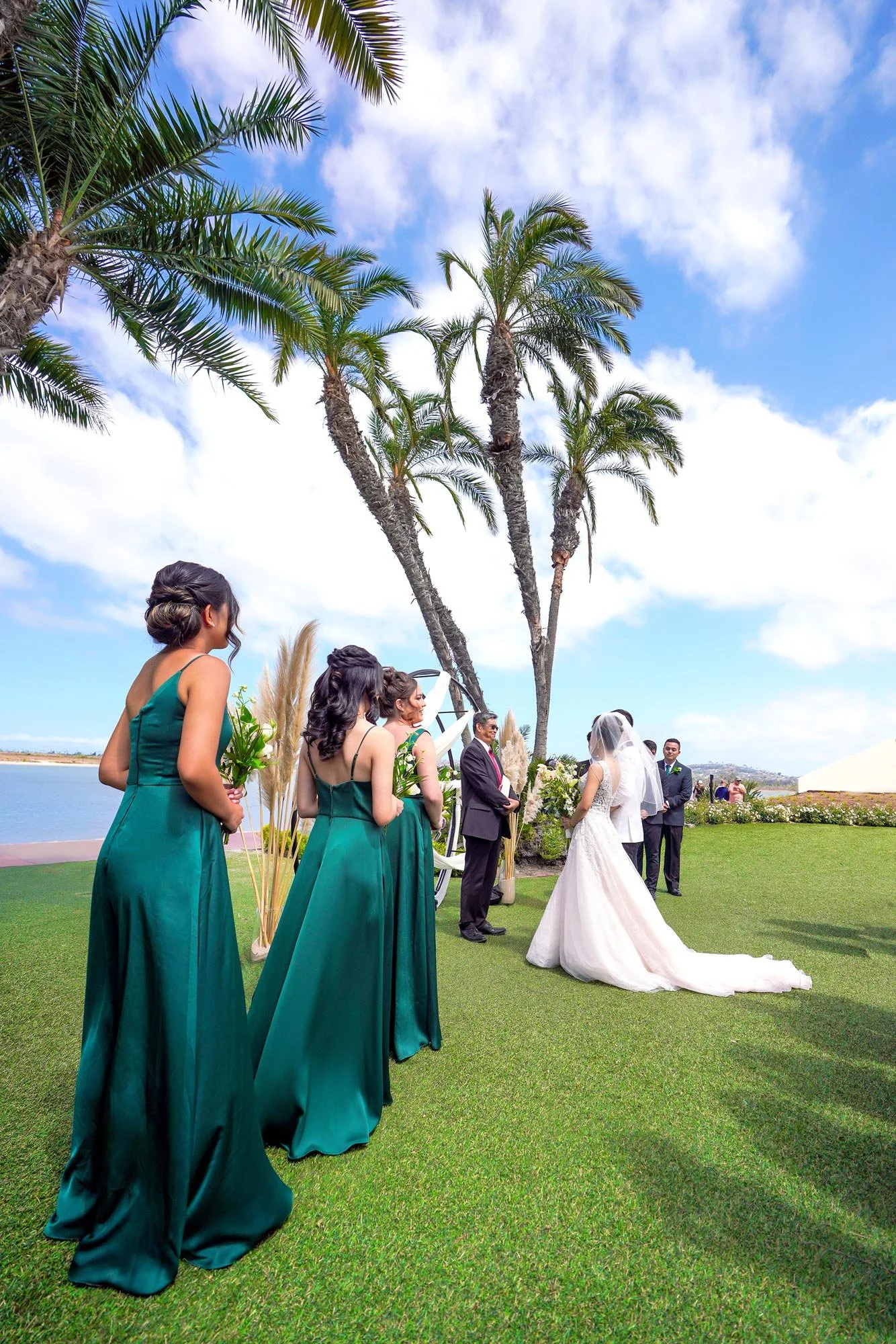 A view from behind three bridesmaids in emerald green gowns, looking towards the bride and groom standing at the altar during a sunny outdoor wedding ceremony on a green lawn with tall palm trees and a body of water in the background at Mission Bay, 