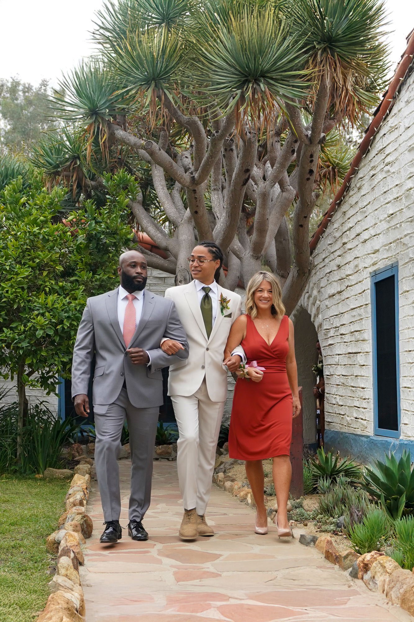 A photo of groom with his parents walking down an outdoor flagstone path: a man in a grey suit, a man in a cream suit and olive tie, and a woman in a reddish-orange dress, with a unique large tree and white shingled building in the background.