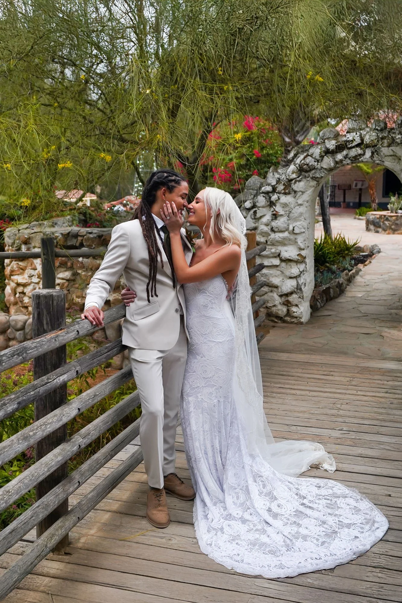  photo of a newly married couple embracing and about to kiss on a wooden bridge, with one bride wearing a cream-colored suit and the other in a white lace wedding dress with a long train and veil. A stone archway and lush green foliage with pink flow