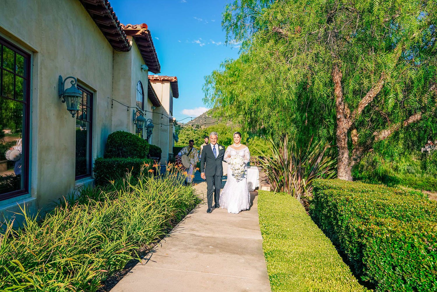 A daytime outdoor photograph of a bride in a white long-sleeved lace wedding dress being escorted down a paved walkway by an older man in a dark suit. The walkway is lined with green hedges and plants. They are walking beside a beige stucco building 