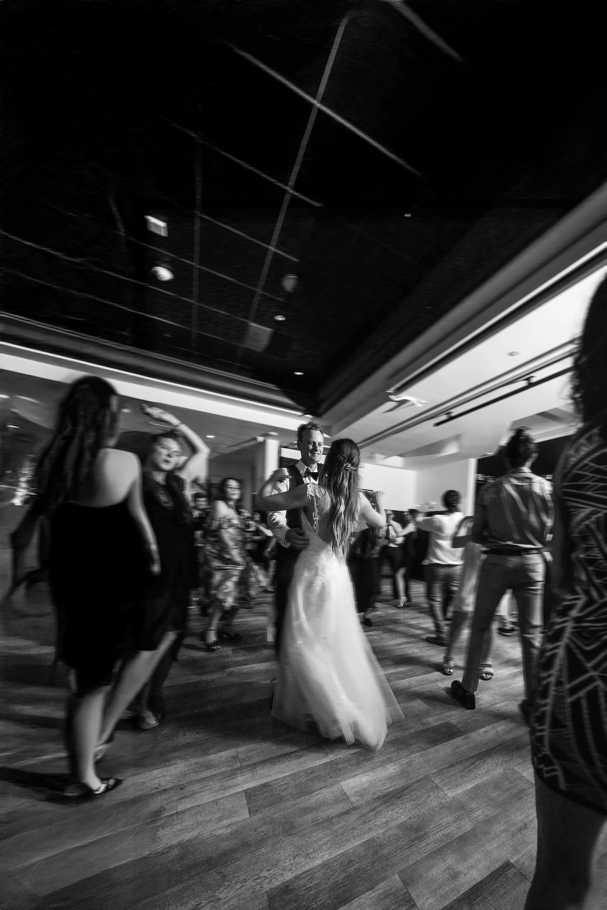 A black and white, low-angle shot captures a lively wedding reception on a wooden dance floor. In the center, a bride in a backless white gown and a groom in a dark suit share a dance, surrounded by guests in motion. To the left, a woman in a dark dr
