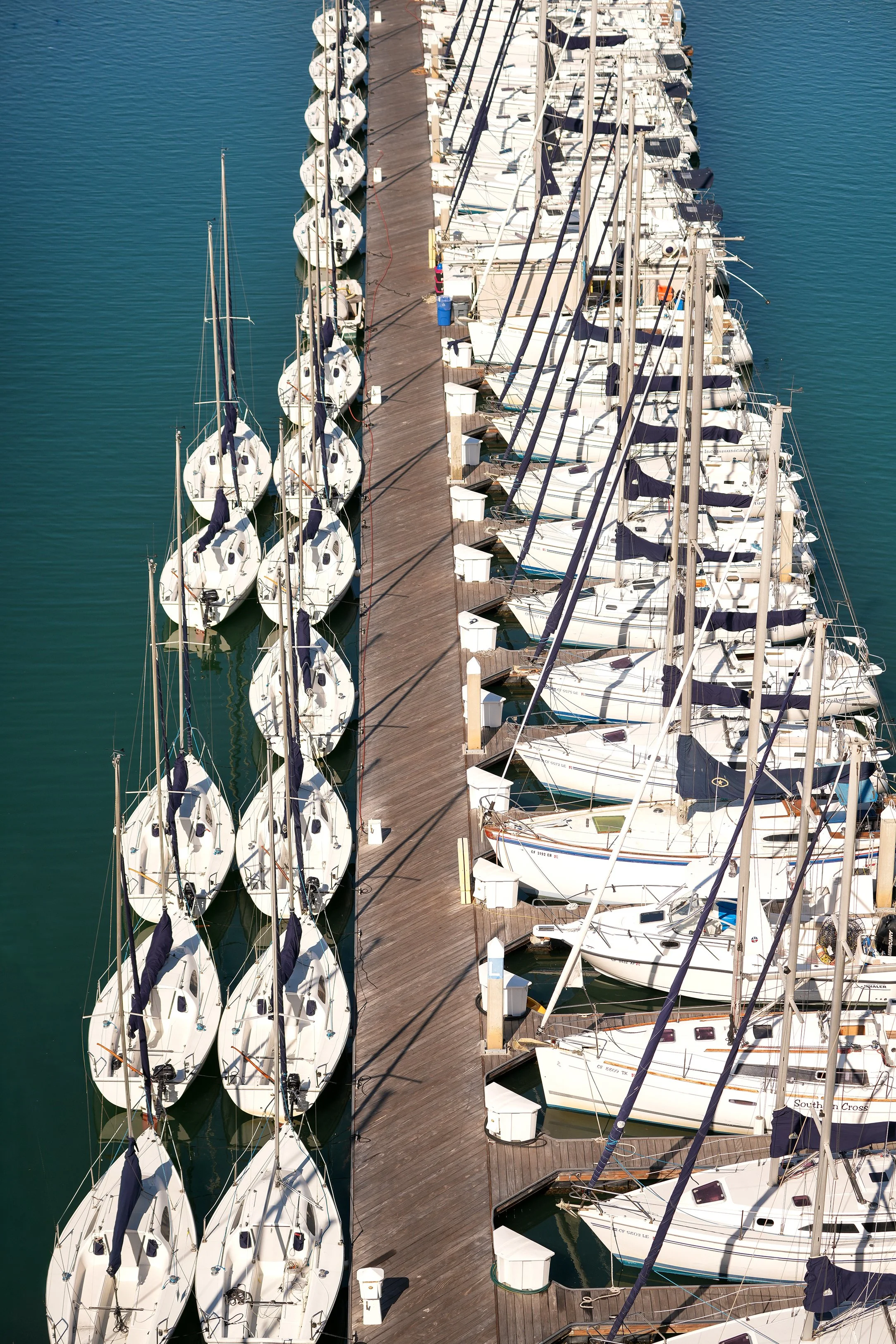 An elevated view of a marina showing two rows of white sailboats docked along a wooden pier, with clear blue water and strong shadows.