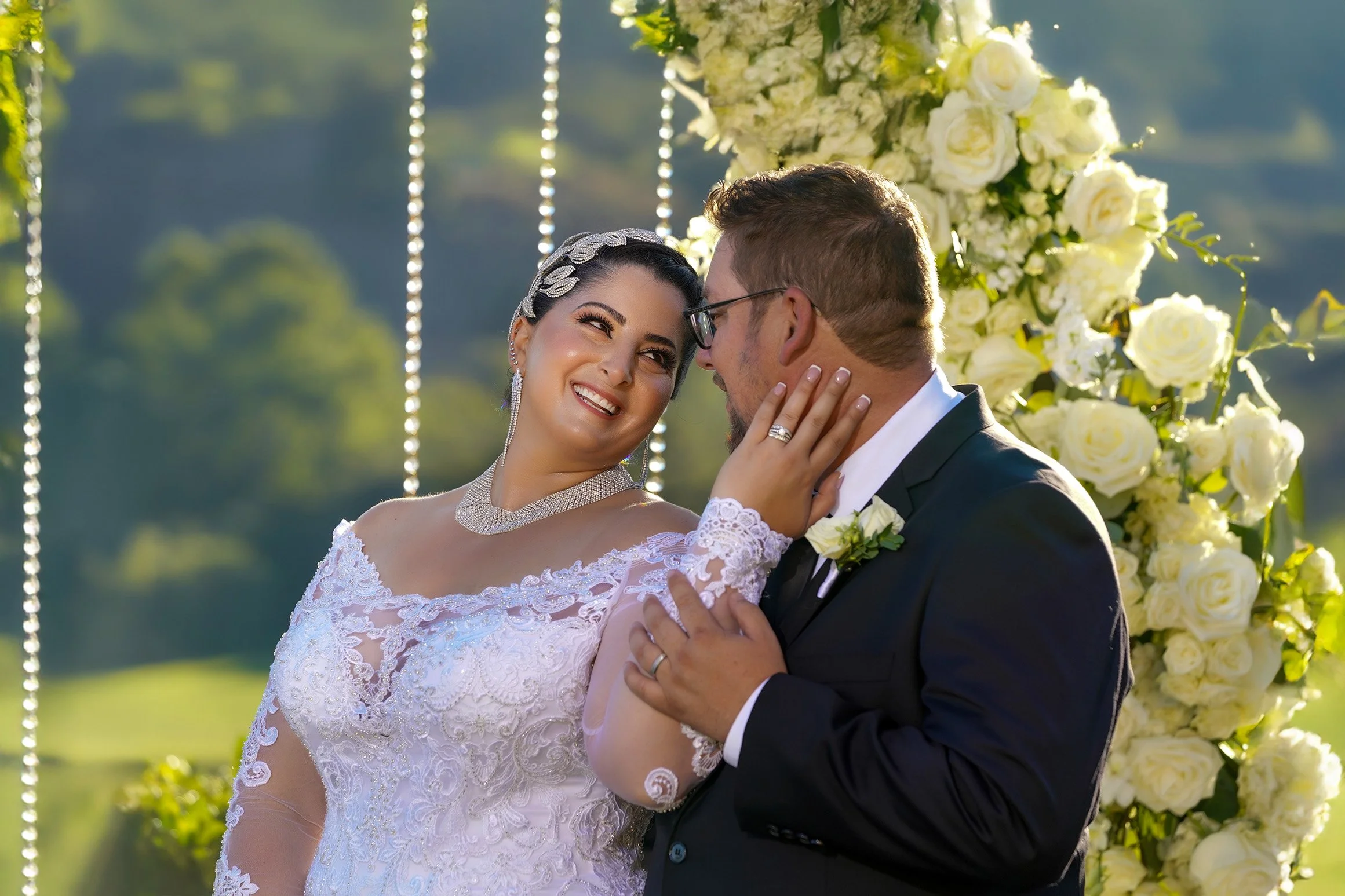 A smiling bride and groom share an intimate moment outdoors under a floral arch of white roses. The bride, in an off-the-shoulder lace gown, jeweled headpiece, and necklace, gazes at the groom while resting her hand on his cheek. The groom, wearing a