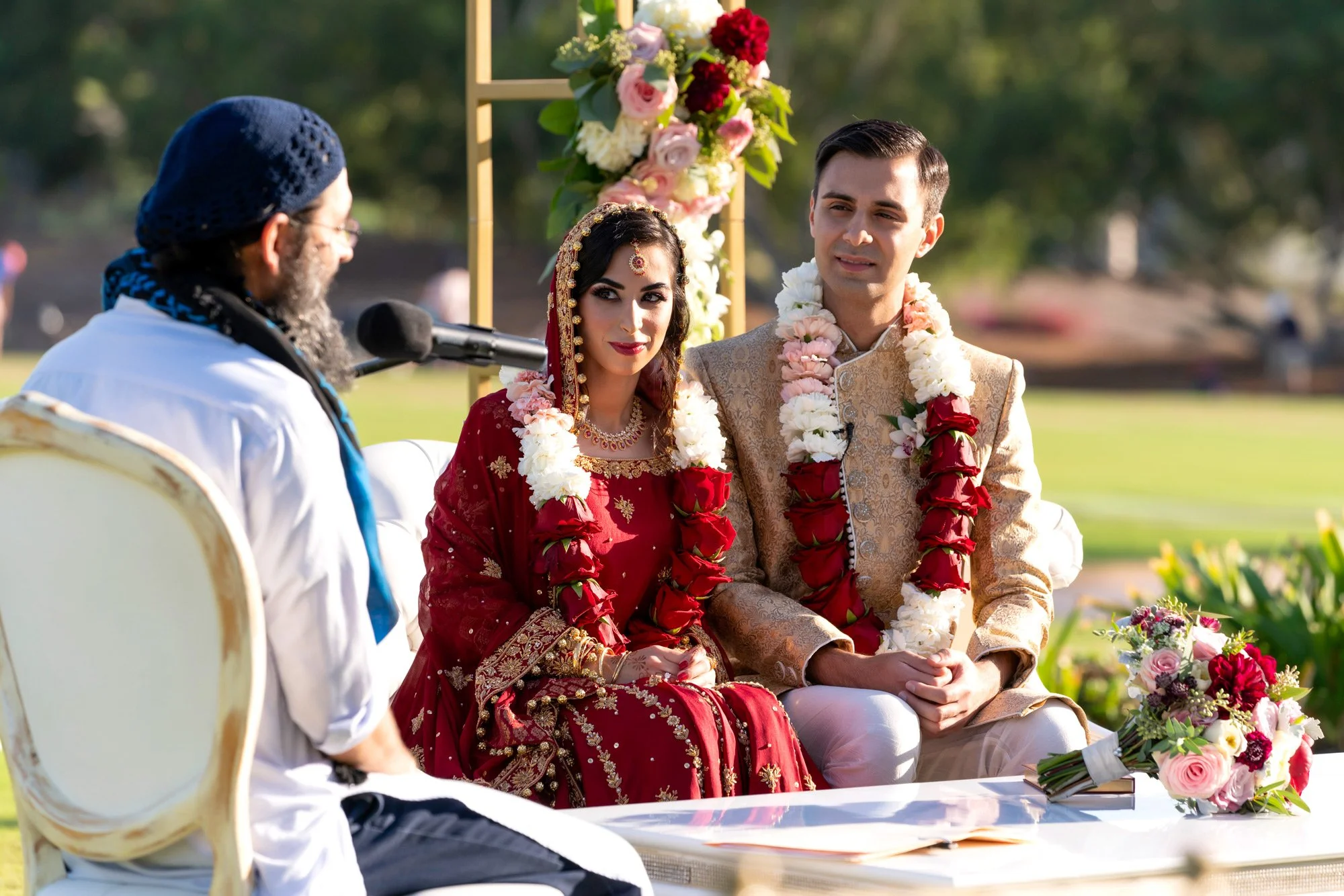 An outdoor photograph of a bride and groom seated at their wedding altar during a ceremony. The bride wears a traditional deep red lehenga with gold accents and a veil, while the groom wears a gold sherwani. Both are adorned with red and white flower