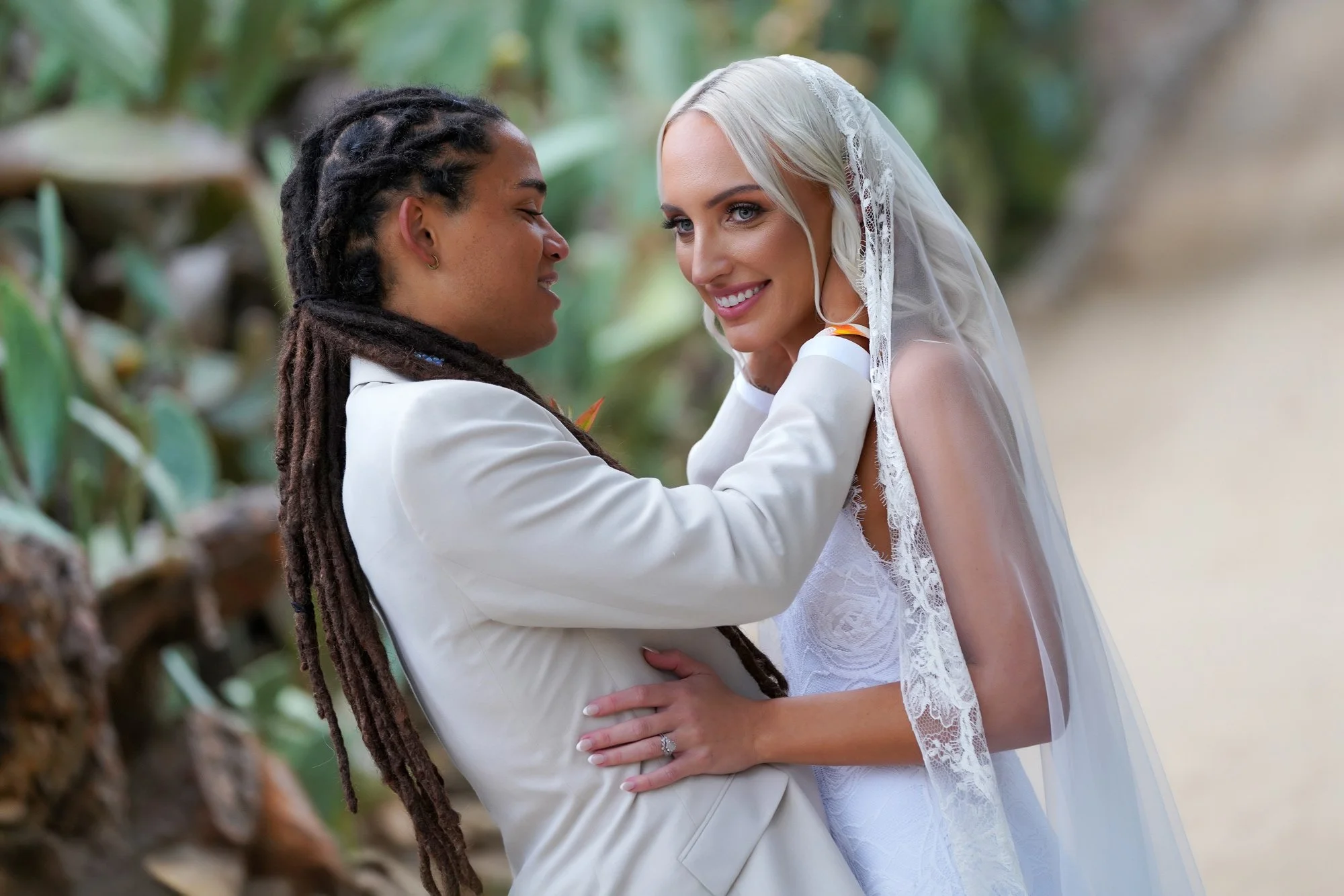 A photo of two brides embracing and smiling lovingly at each other during their outdoor wedding ceremony, with one wearing a cream-colored suit and the other in a white lace wedding dress with a lace-trimmed veil.