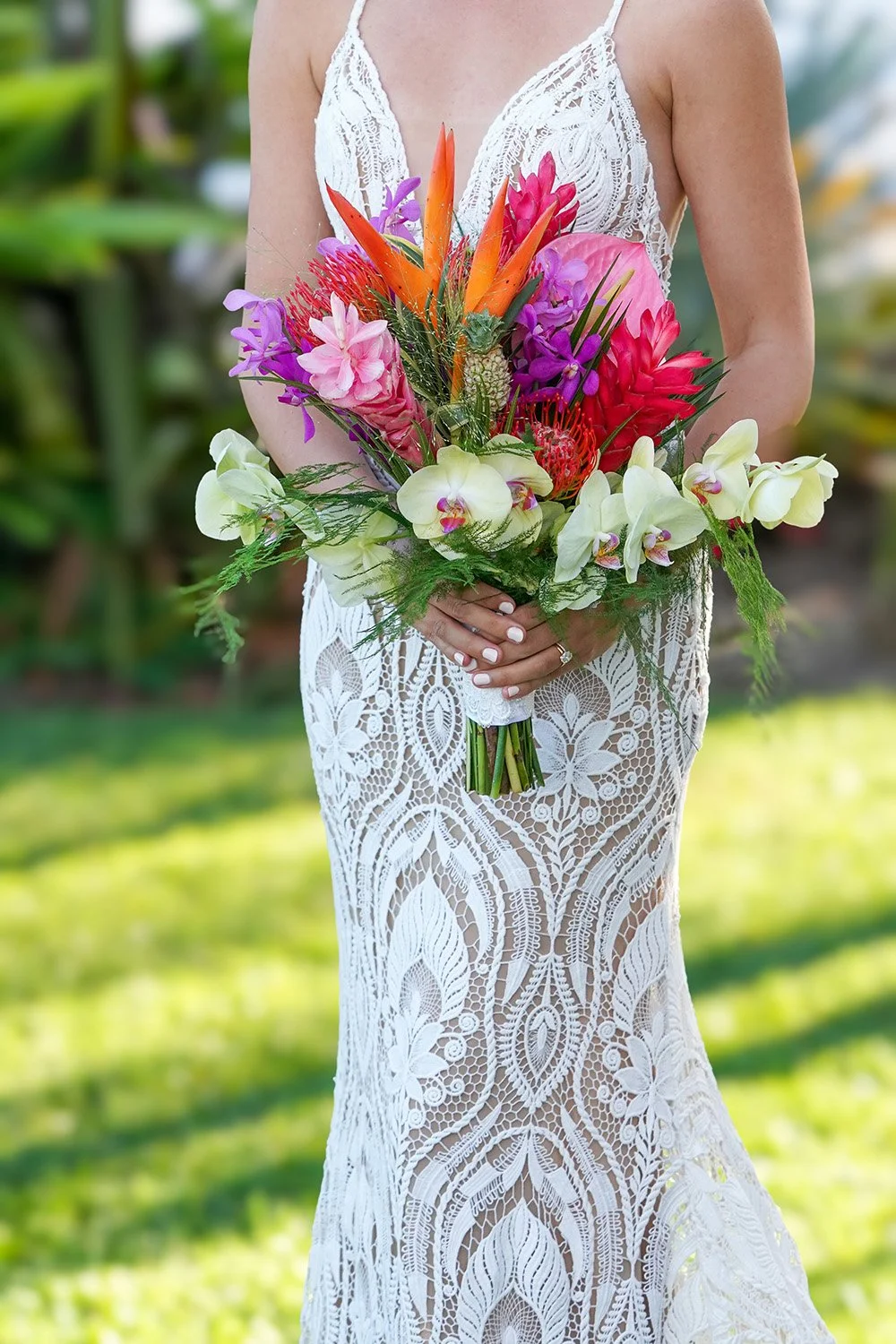 A close-up of a person in a white lace dress holding a large, vibrant tropical wedding bouquet featuring yellow orchids, orange heliconia, pink ginger, and other assorted bright flowers.
