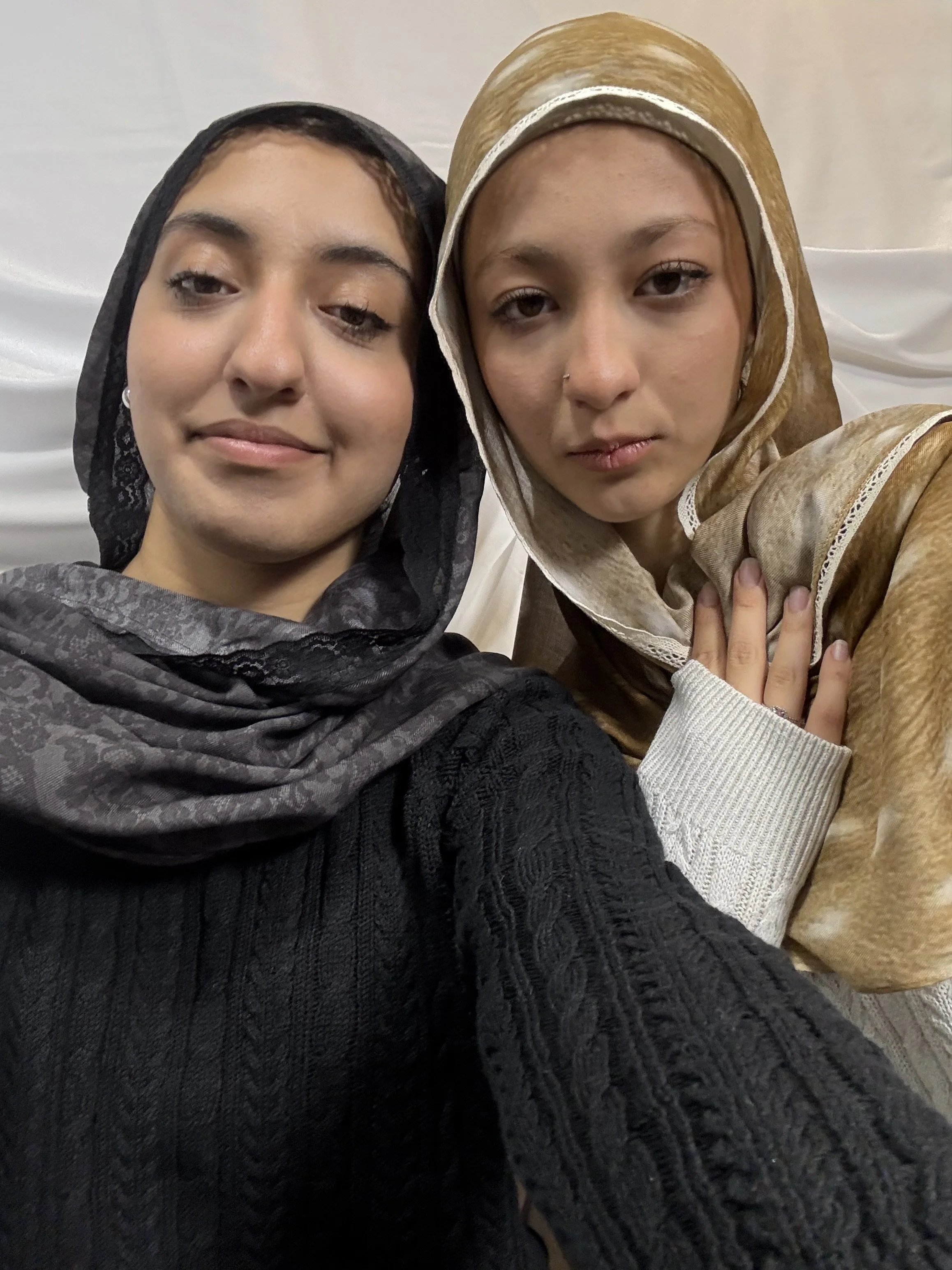 Two women wearing headscarves taking a close-up selfie against a plain background.
