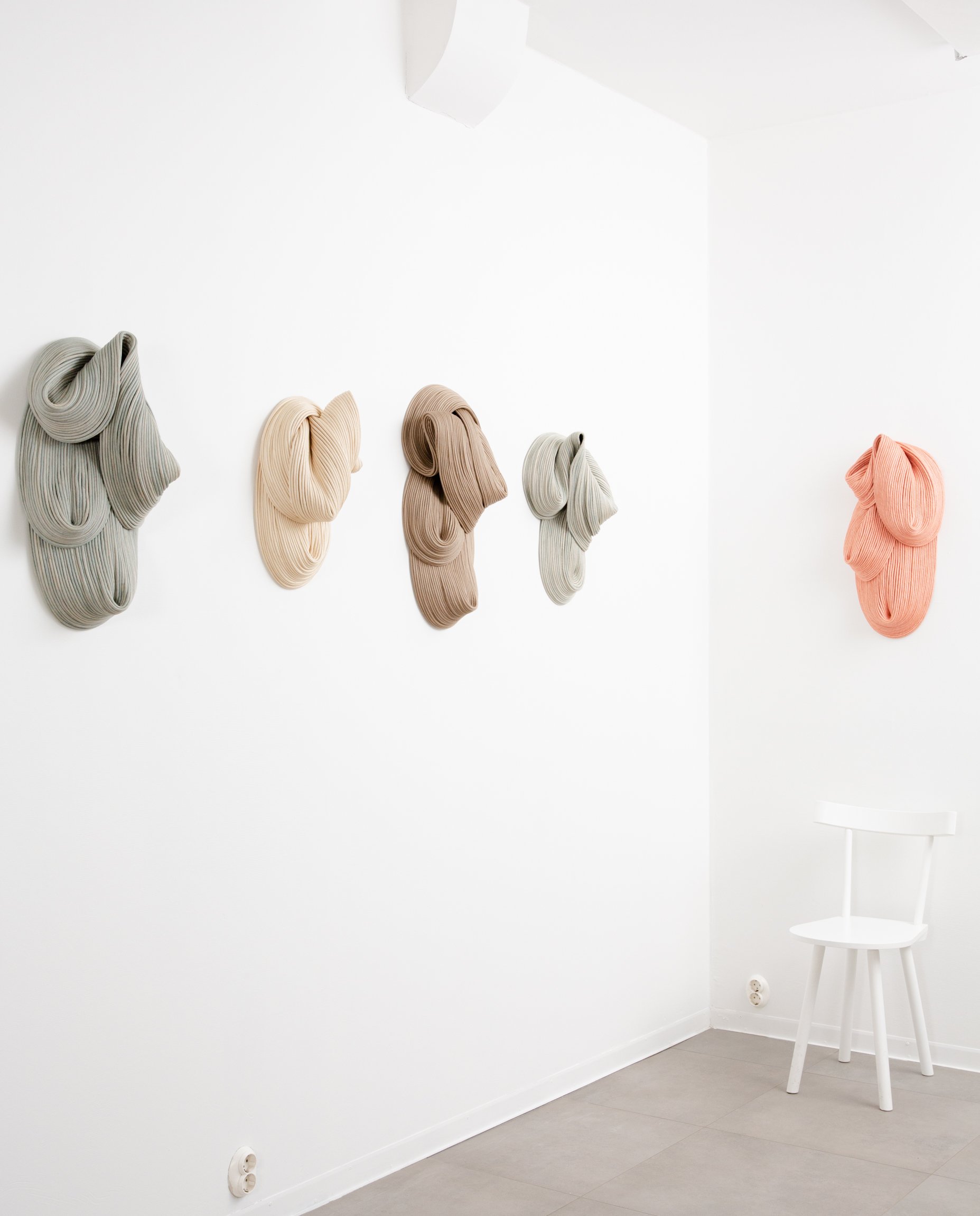 Colorful woven wall hangings in various shades of beige, gray, brown, and peach arranged on a white wall in a minimalistic room with a white chair underneath the peach wall hanging.