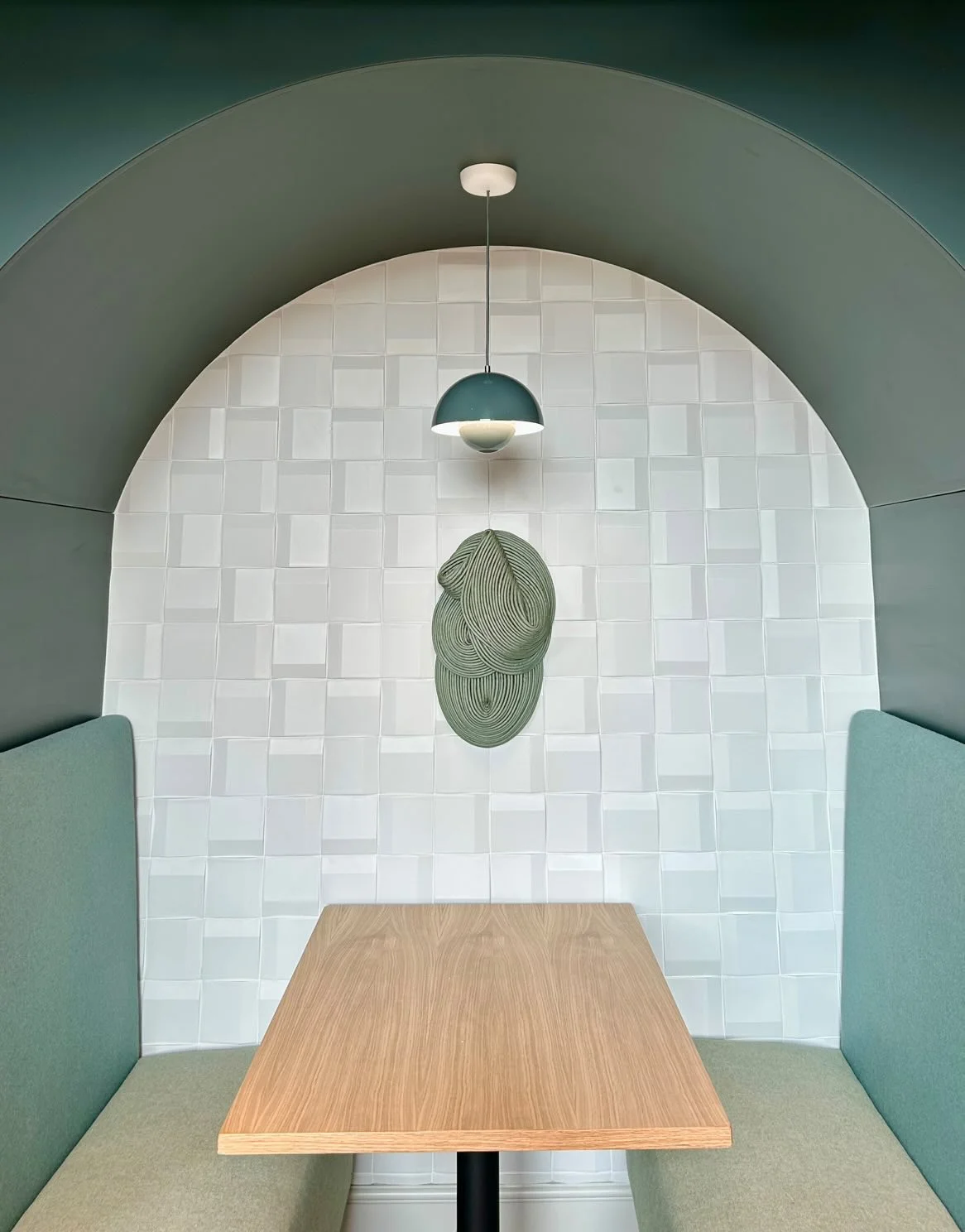A small booth with seating on either side of a wooden table, a textured wall with a central artistic wall hanging, and a ceiling light fixture above.