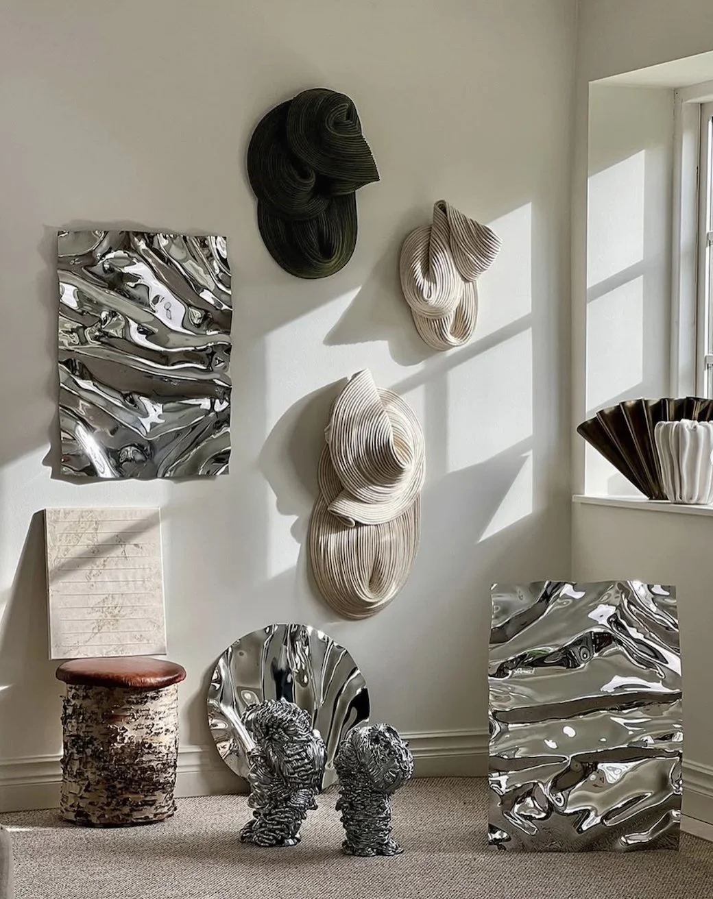 Decorative modern art pieces including metallic sculptures, textured wall hangings, and a small table with a textured surface and ceramic sculptures, displayed against a white wall with sunlight casting shadows.
