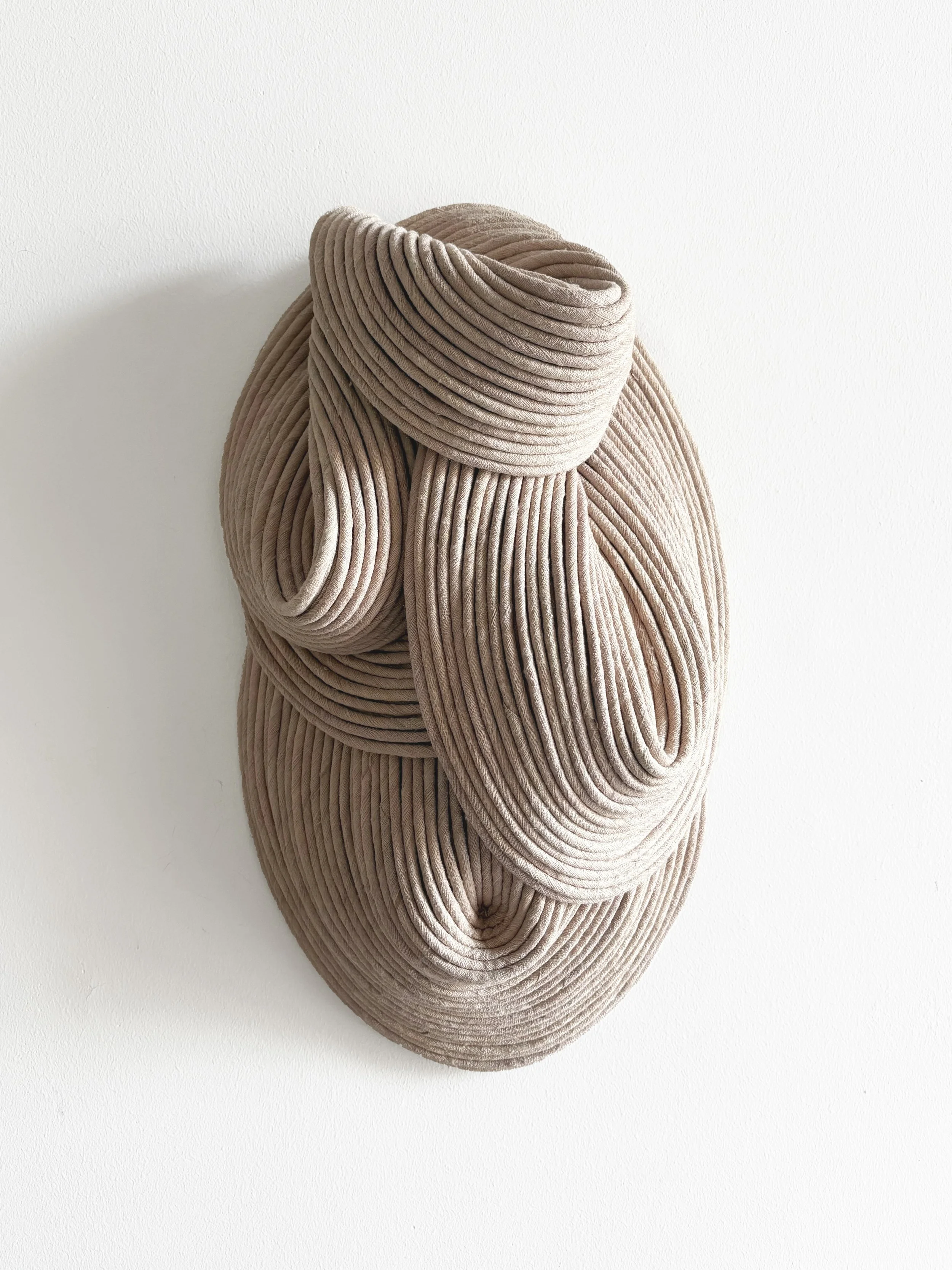 A modern sculpture made of beige, ridged, flexible material, shaped into abstract, flowing curves, mounted on a white wall.