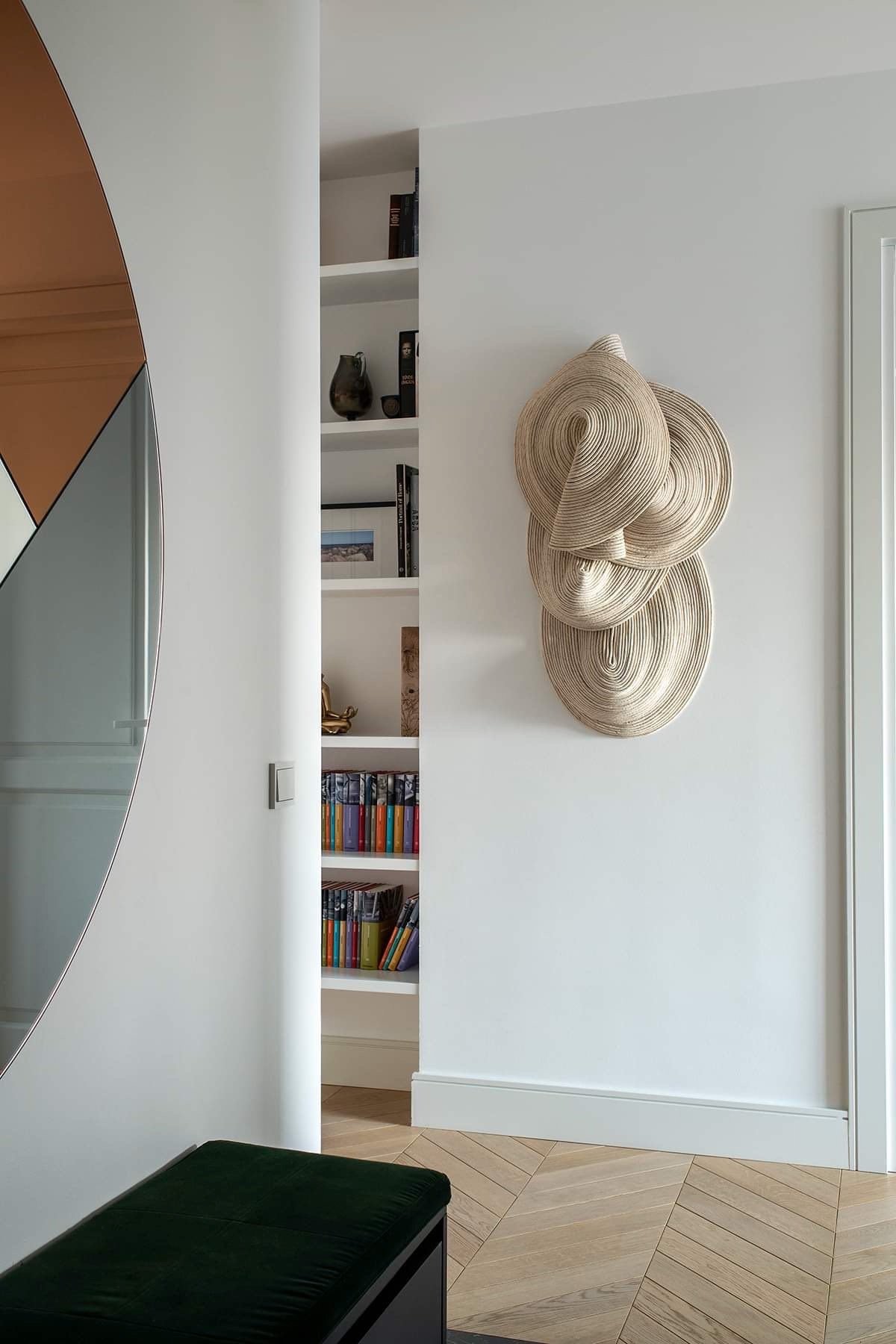 Interior view of a white living space with a built-in bookshelf, decorative beige wall art, and a green bench.