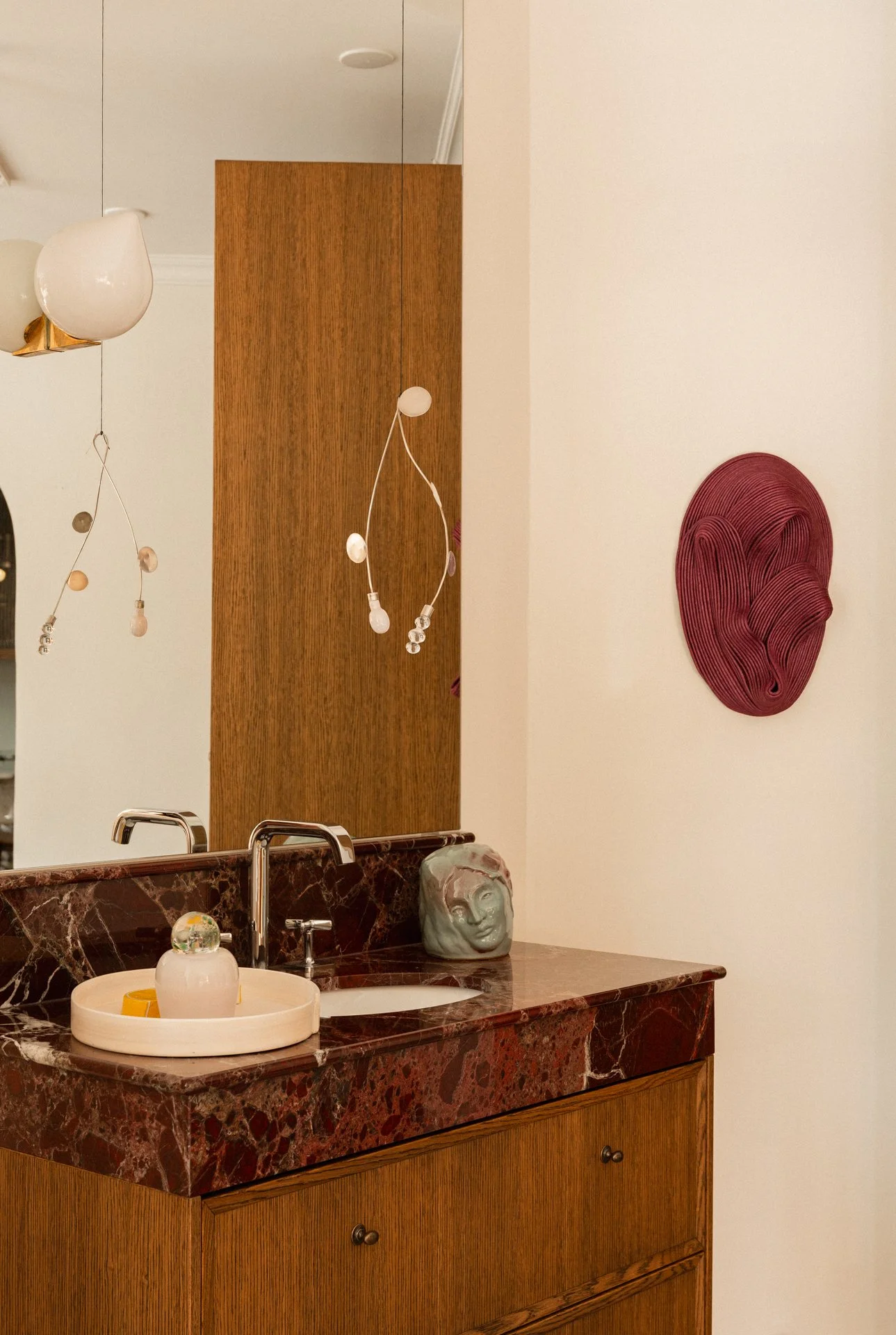 A bathroom vanity with a maroon marble countertop, a wooden cabinet, and a small ceramic head sculpture on top. There is a glass orb and a tray on the sink, decorative hanging light fixtures, and a piece of abstract wall art on the wall.