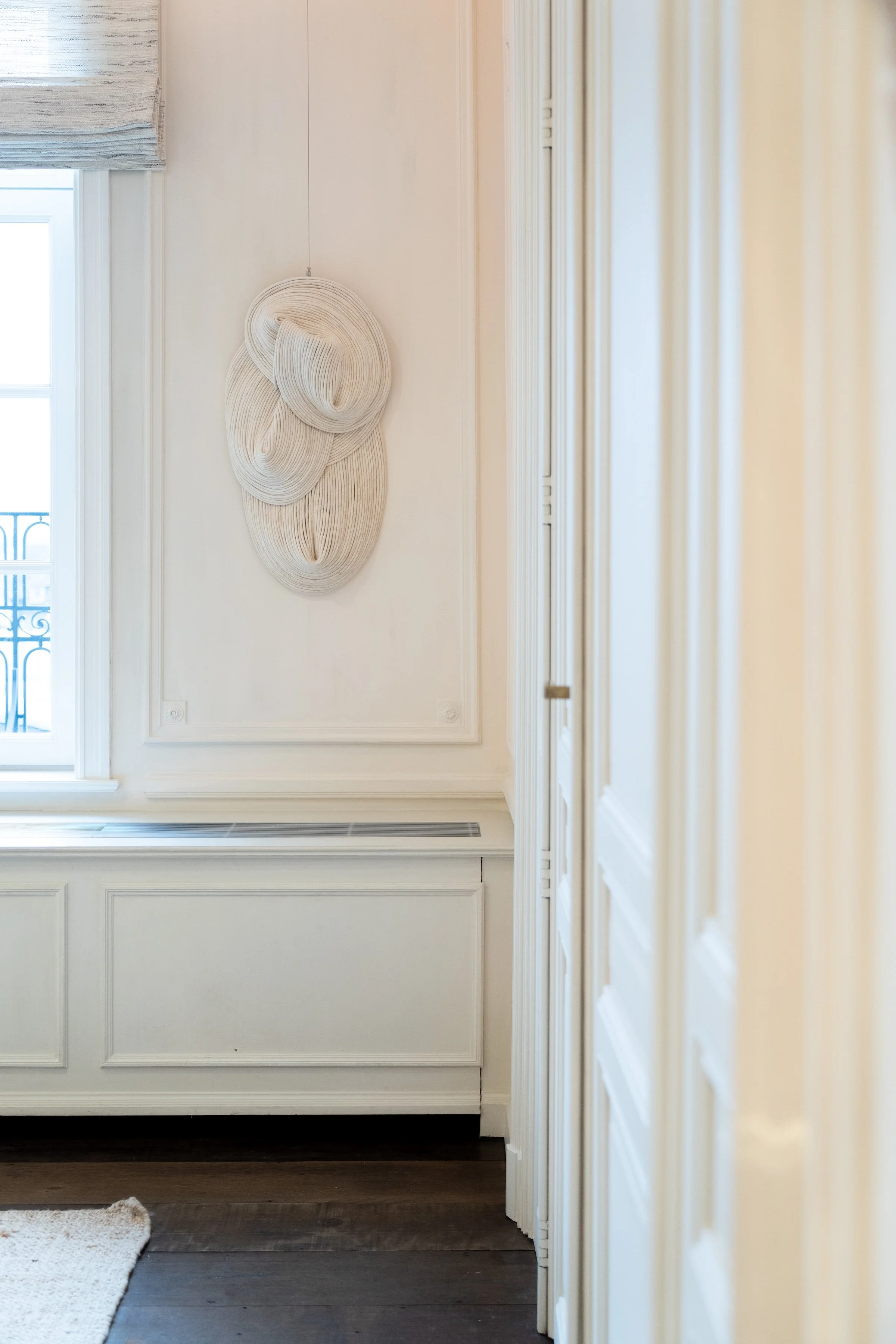 A corner of a room with white paneled walls, a window with a roman shade, white decorative molding, and a woven wall hanging art piece.